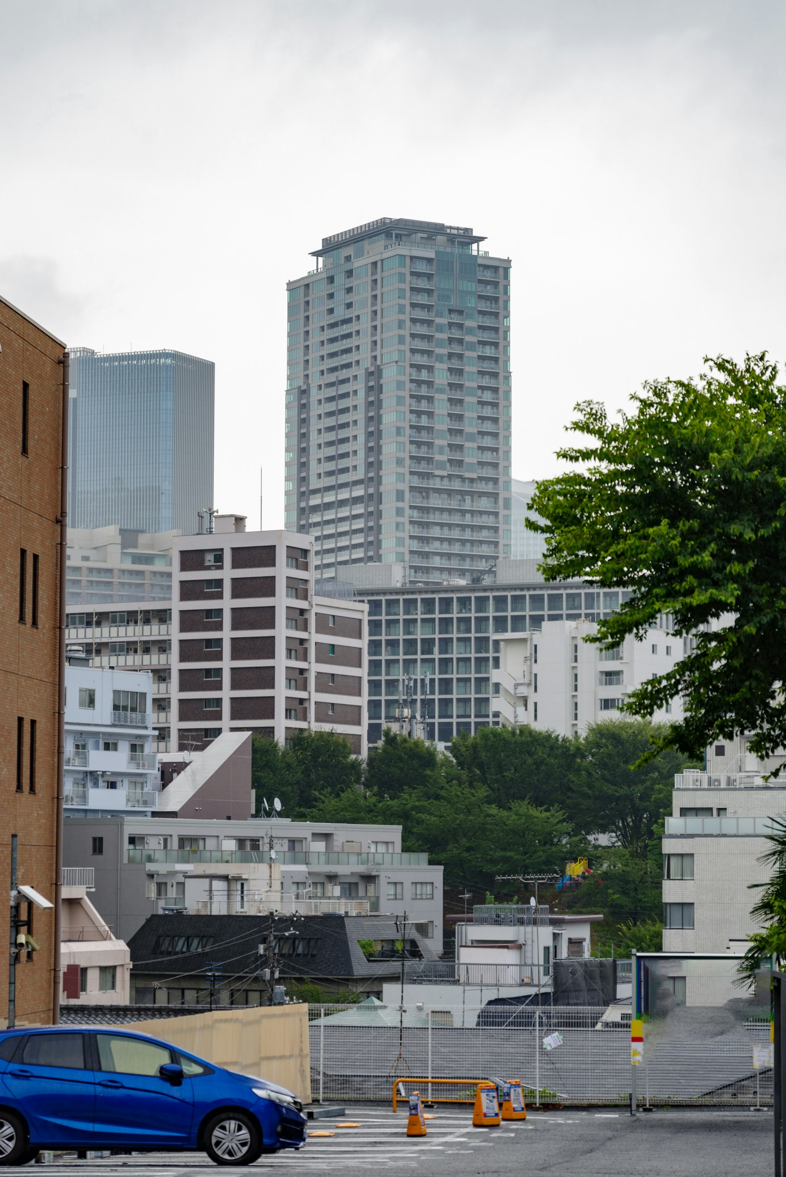 Cityscape featuring tall buildings a blue car in the foreground and green trees on the left