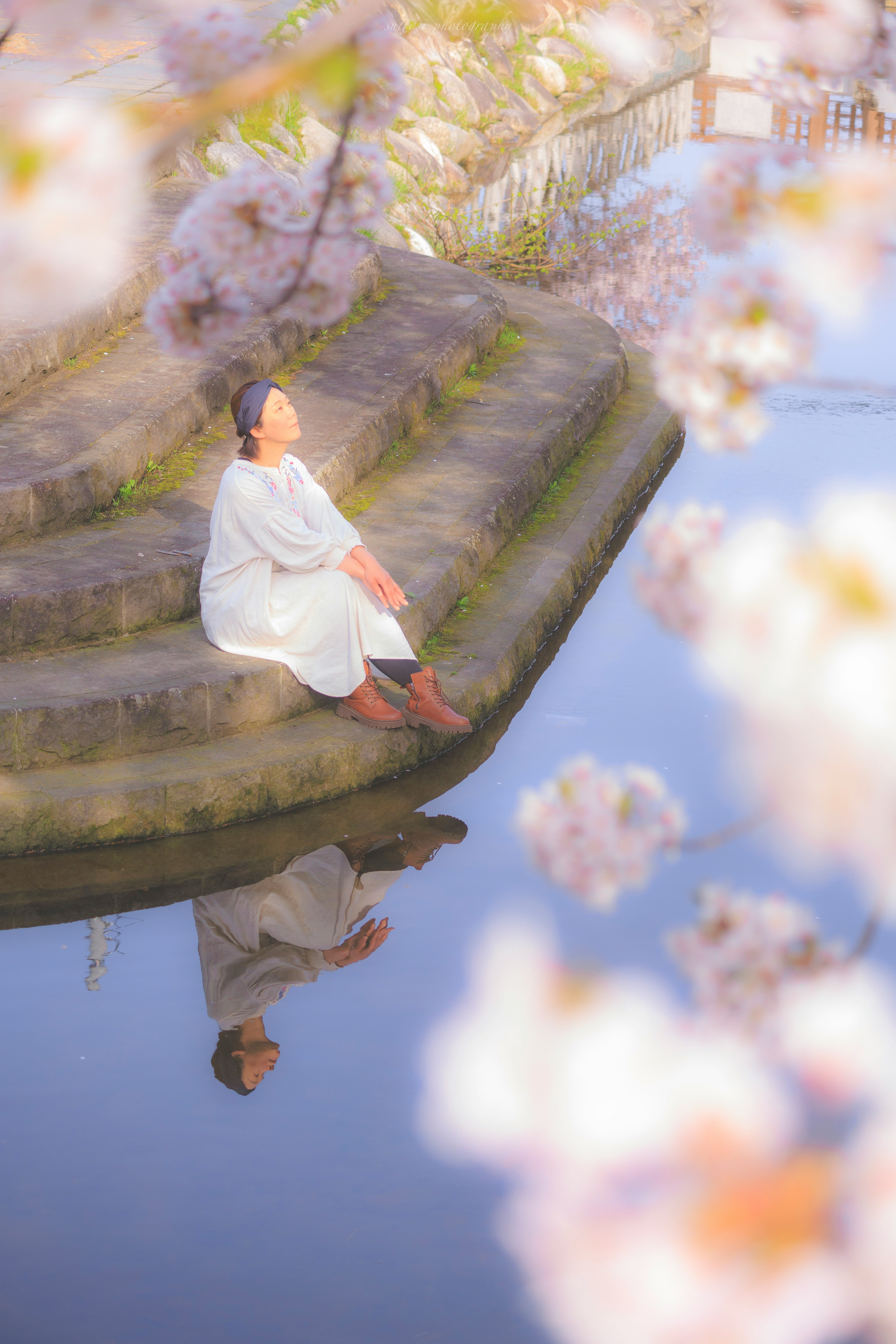 水辺の石段で座る白い衣の人物と桜の花びらのボケ