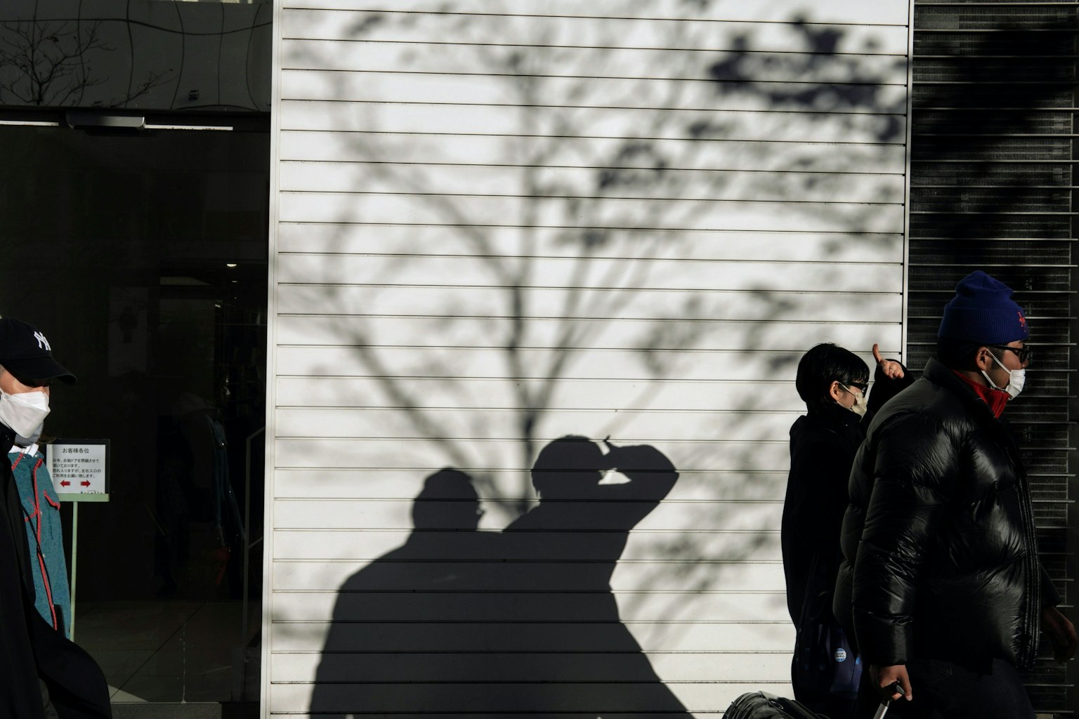 Personas con mascarillas caminando con sombras proyectadas en una pared blanca