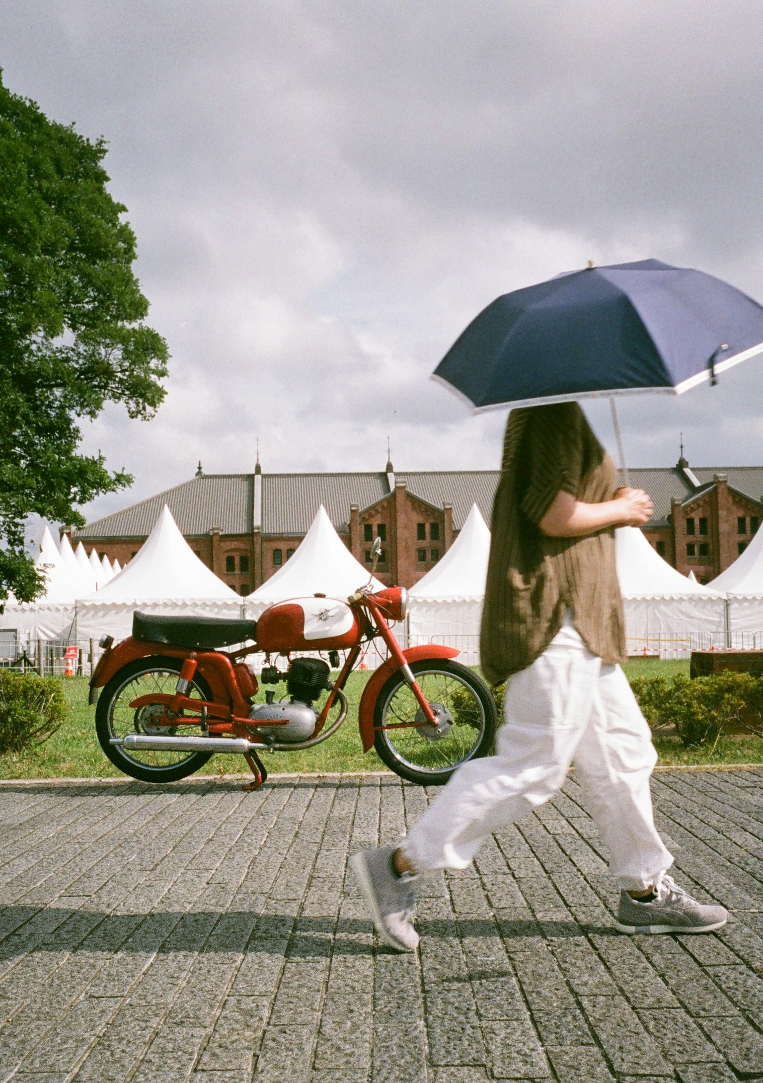 A person holding an umbrella walking past a red motorcycle and white tents