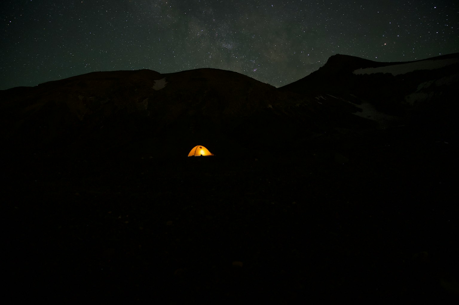 Una pequeña tienda brilla en la oscuridad bajo un cielo estrellado con siluetas de montañas
