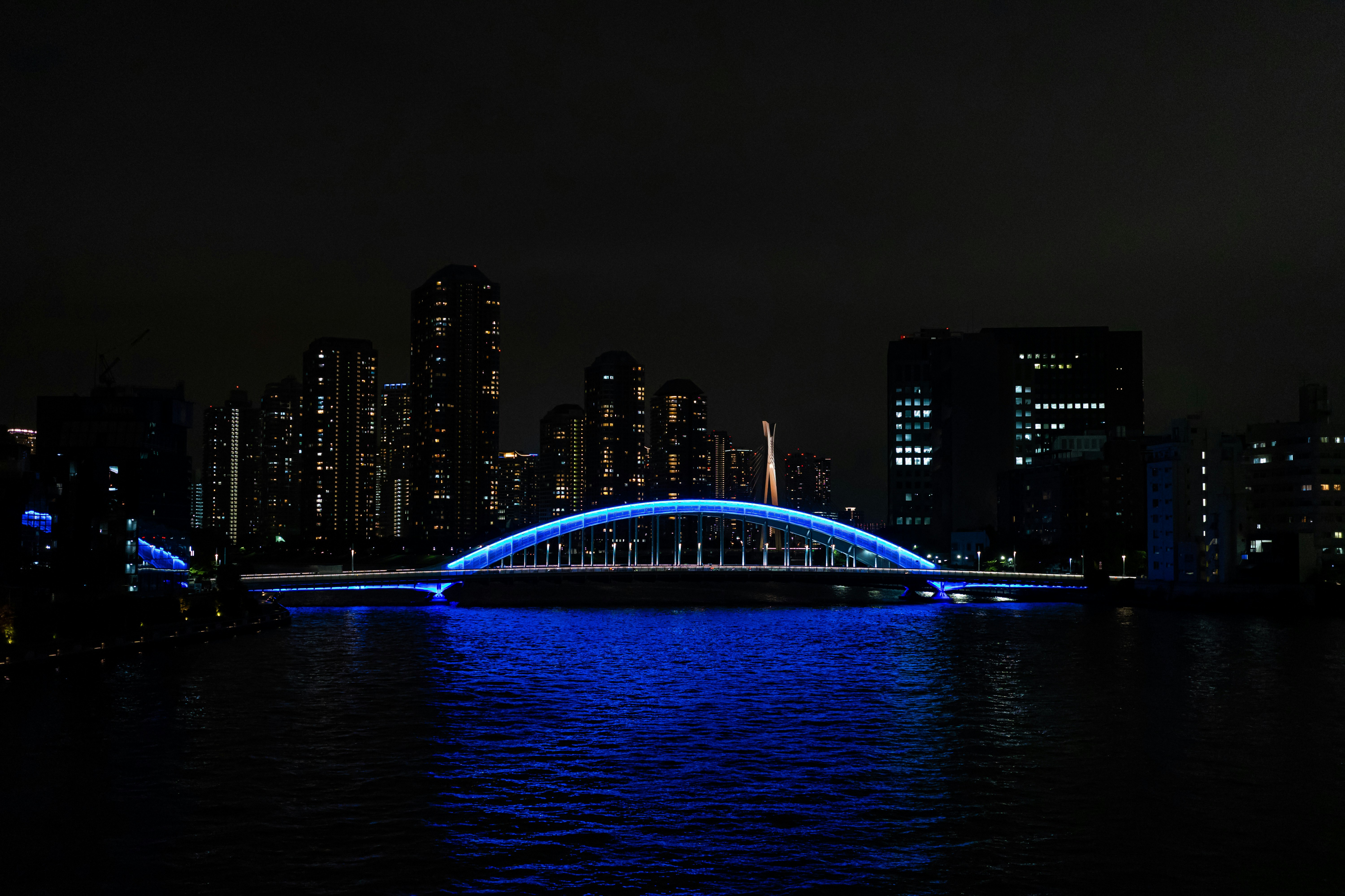 blue-lit bridge over a river at night with city skyscrapers