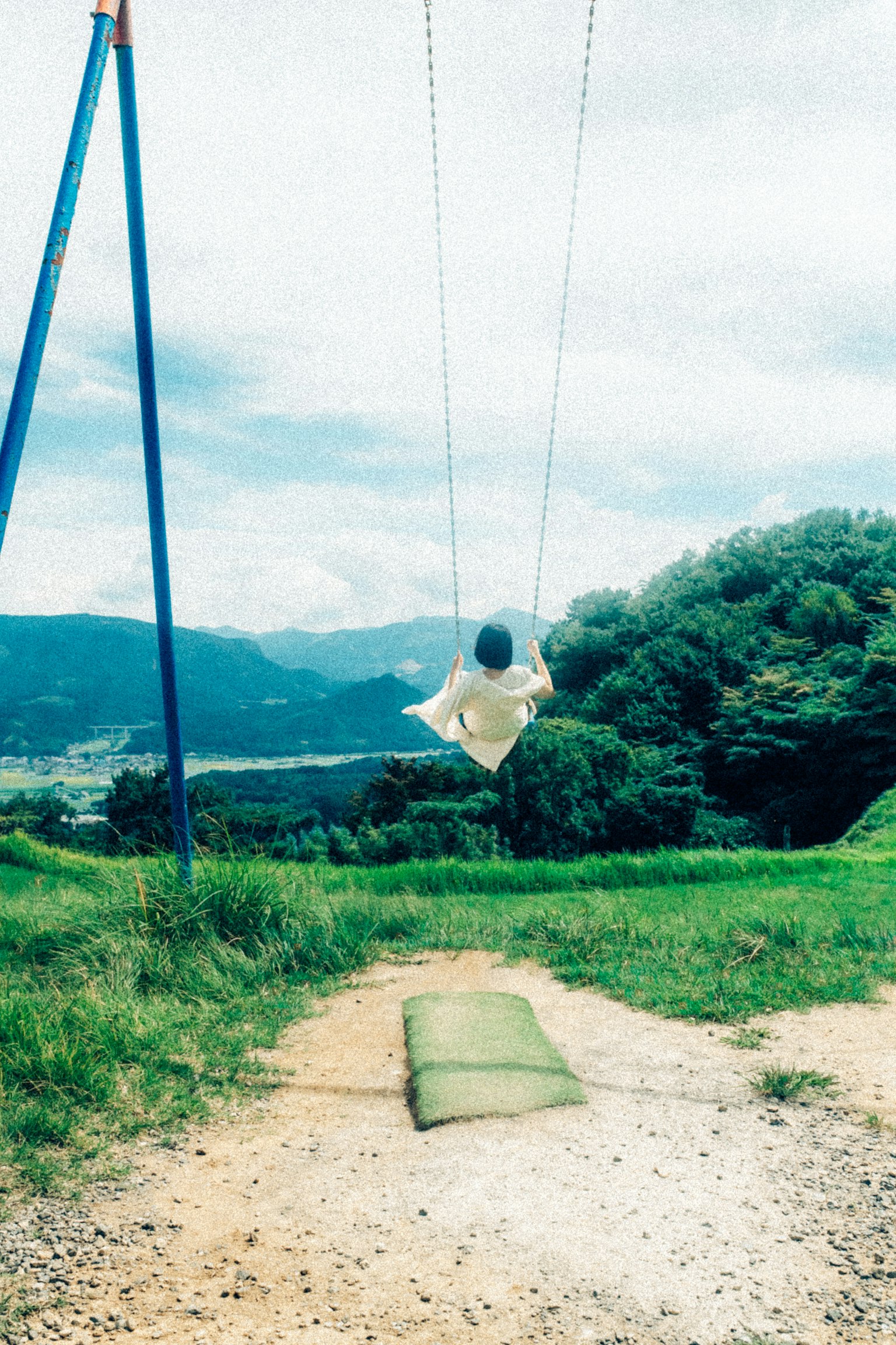 青空の下でブランコに乗る子供の後ろ姿 緑の草原と山々の景色