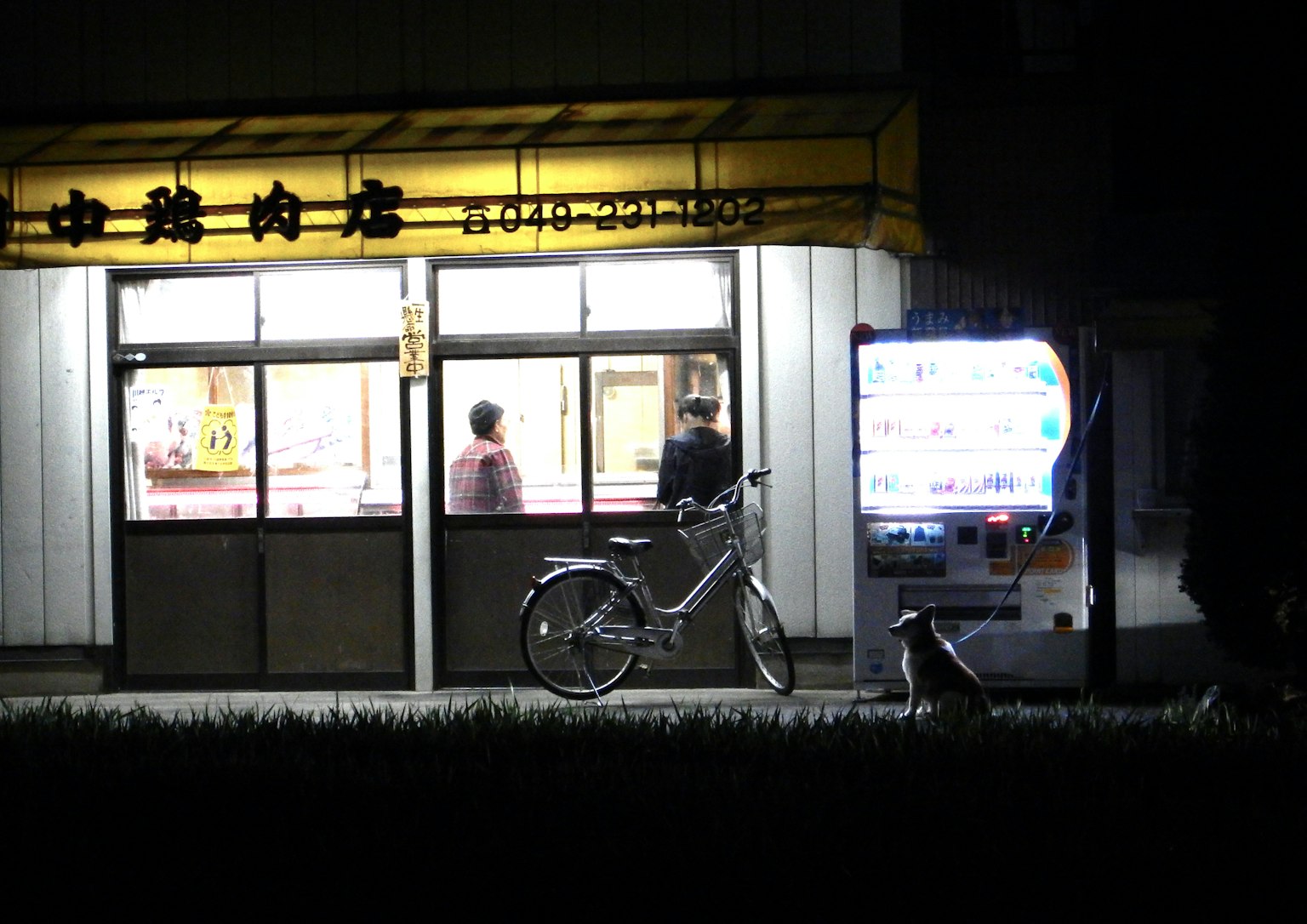ด้านนอกของร้านอาหารในเวลากลางคืนมีจักรยานและตู้จำหน่ายสินค้าอัตโนมัติ