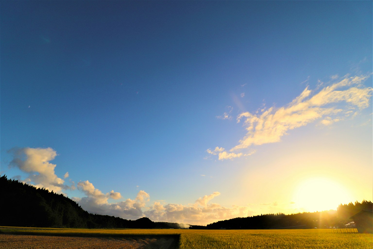 Eine ruhige Sonnenuntergangslandschaft mit einem weiten blauen Himmel und Wolken Sonne über Feldern untergehend
