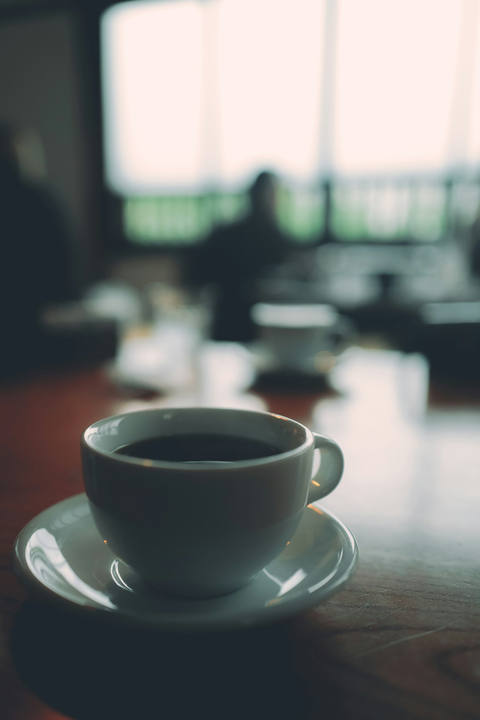 Cangkir kopi dan piring kecil diletakkan di atas meja dalam suasana kafe dengan latar belakang kabur
