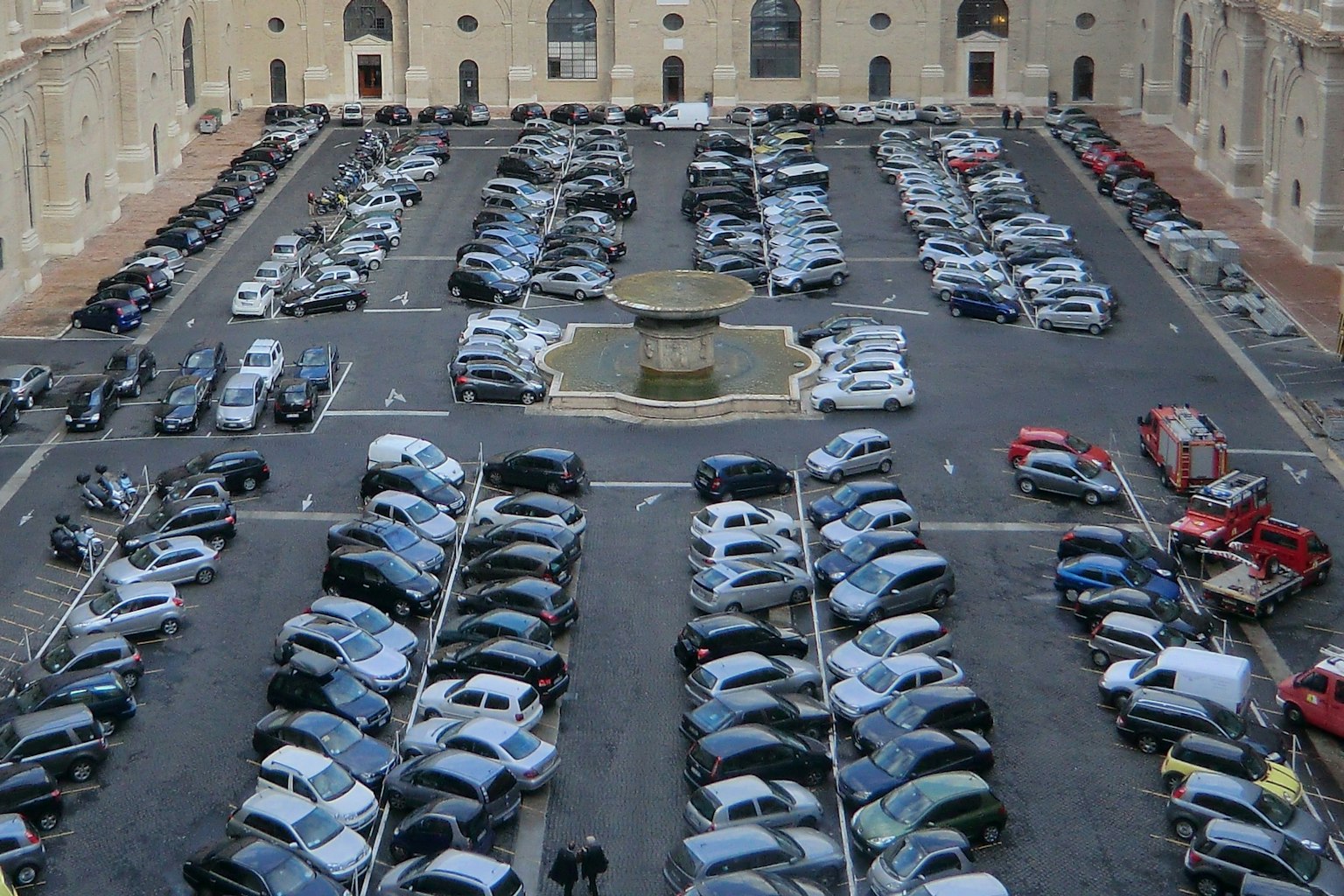 Un grand parking rempli de voitures diverses et une fontaine centrale