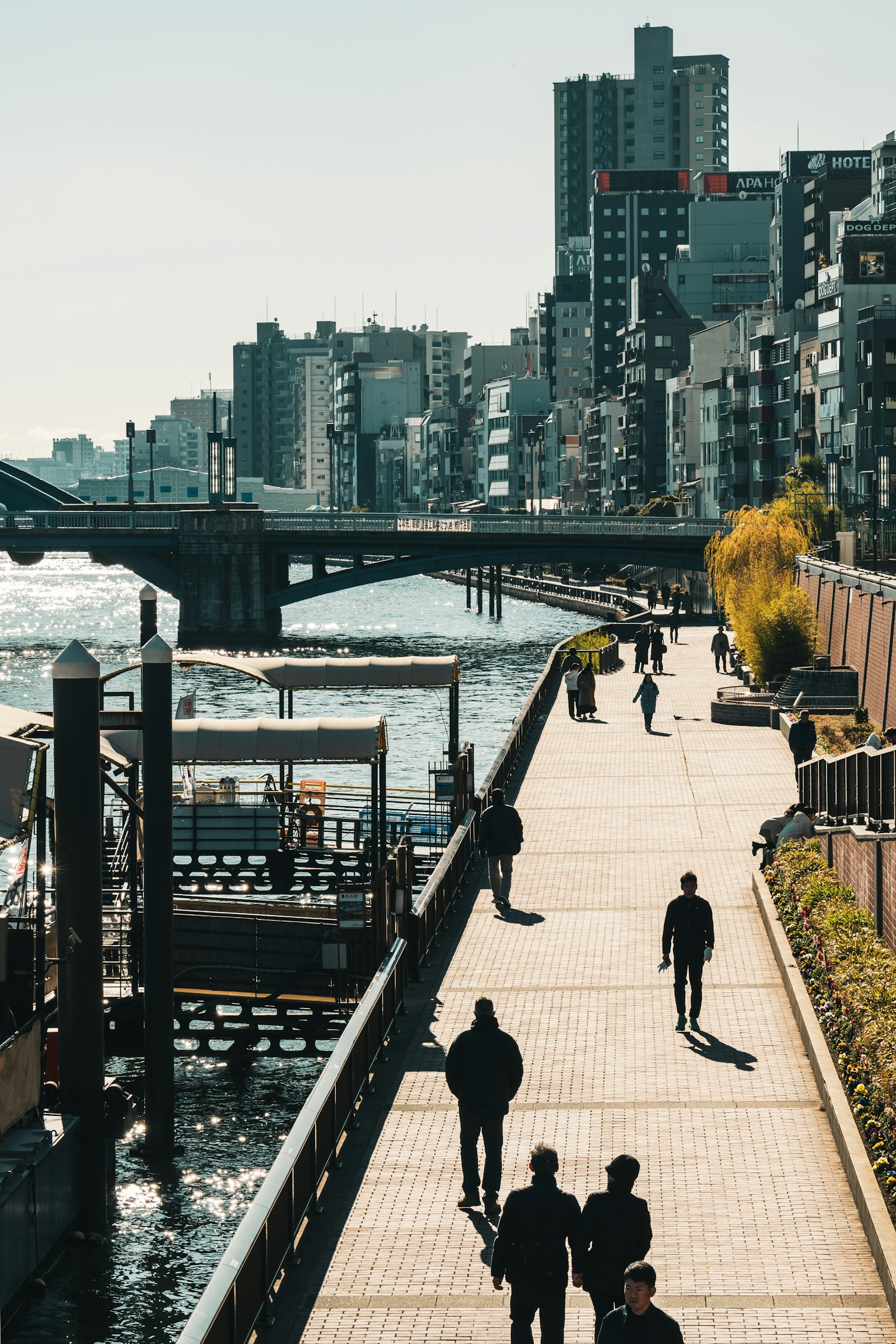 Personas caminando por un paseo junto al río con edificios altos al fondo