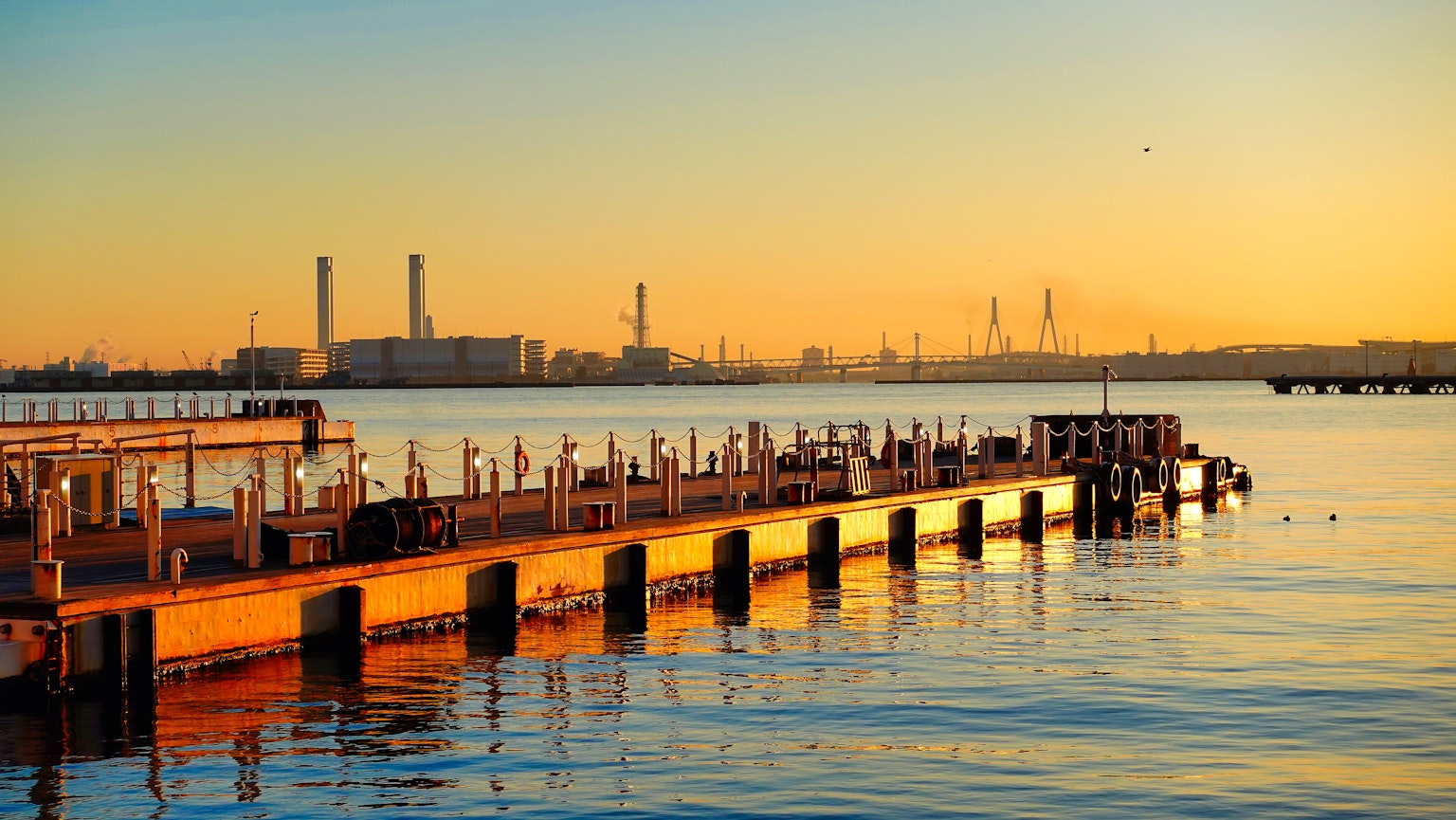 Malersche Aussicht auf einen Steg bei Sonnenuntergang mit Spiegelungen im Wasser und industriellen Schornsteinen im Hintergrund