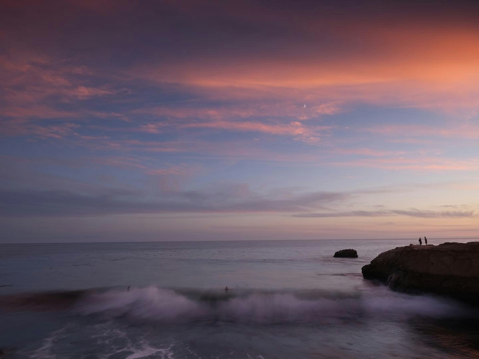 Silhouetten von Menschen an einer felsigen Küste vor einem Sonnenuntergangshimmel