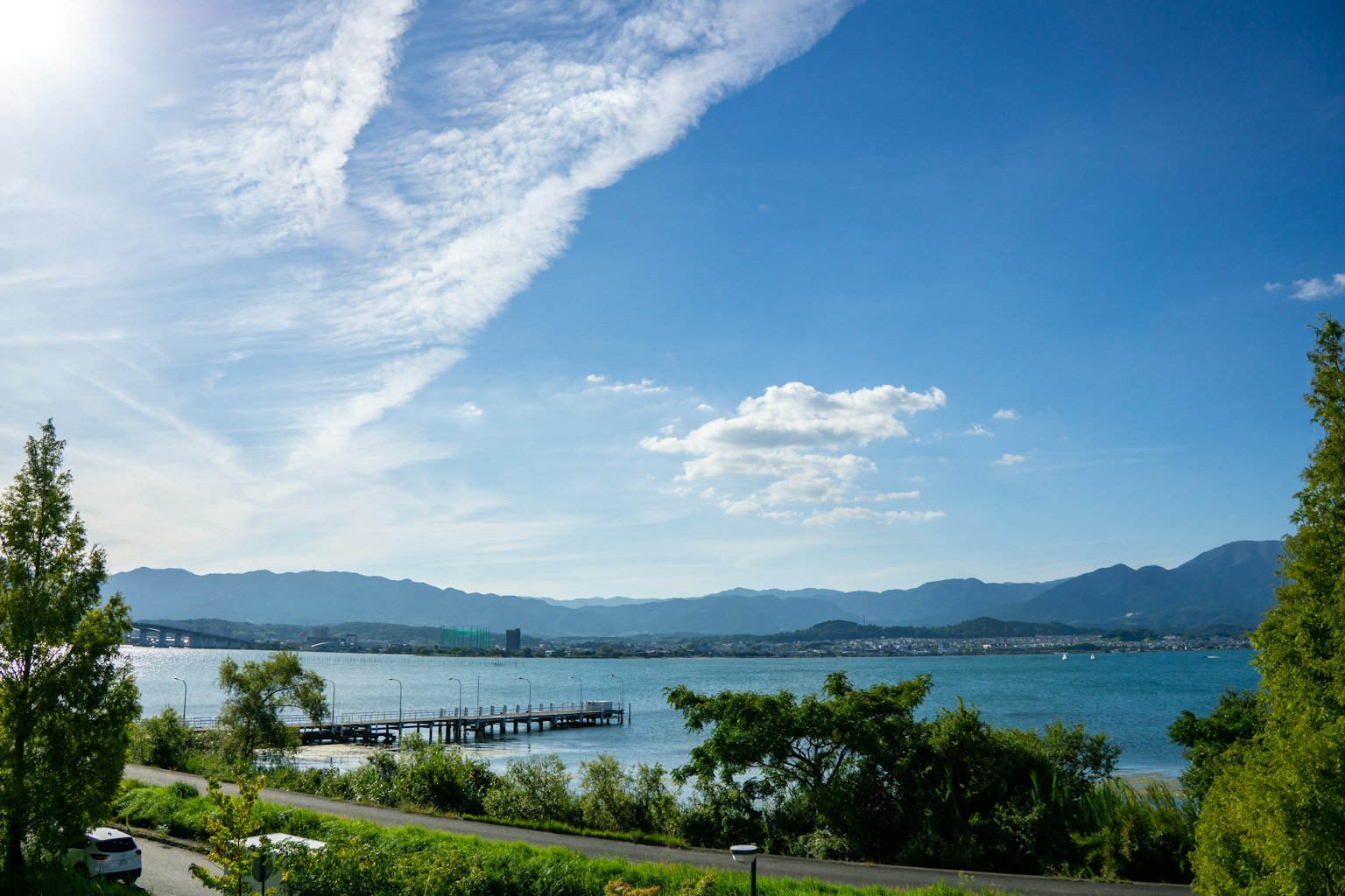 Panoramablick auf einen See unter blauem Himmel mit weißen Wolken grünen Bäumen und einem Steg