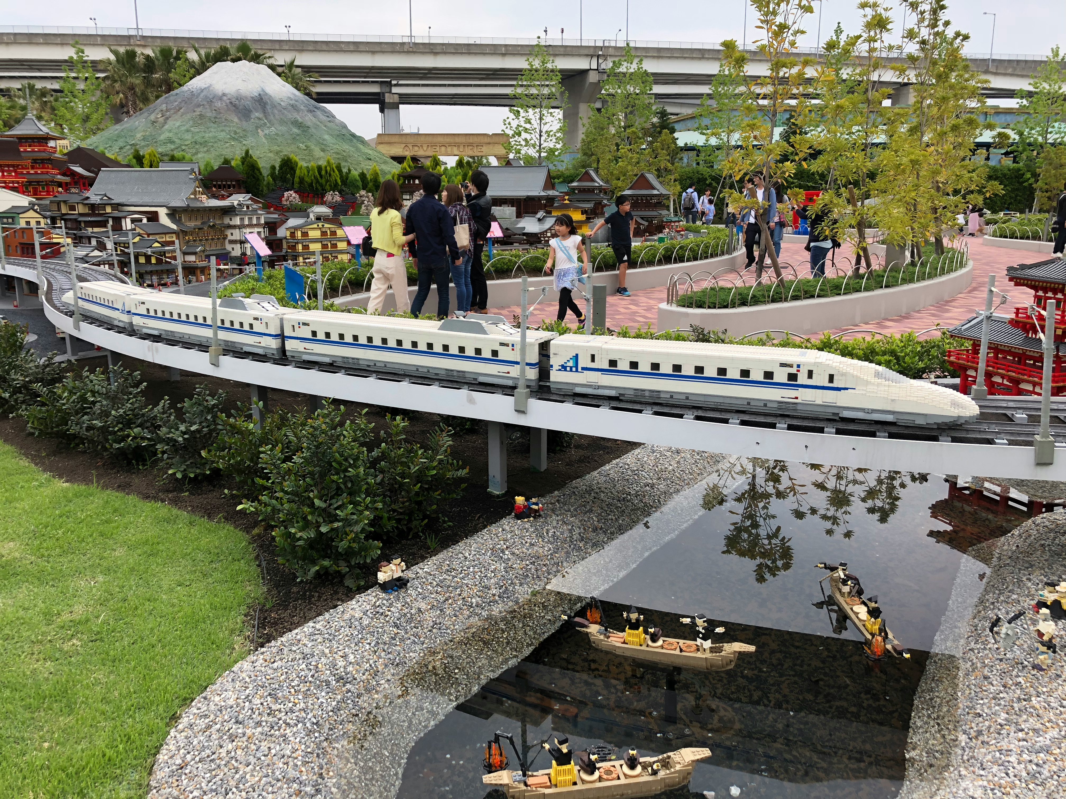 Sebuah kereta Shinkansen miniatur melintasi jembatan di taman dengan pengunjung