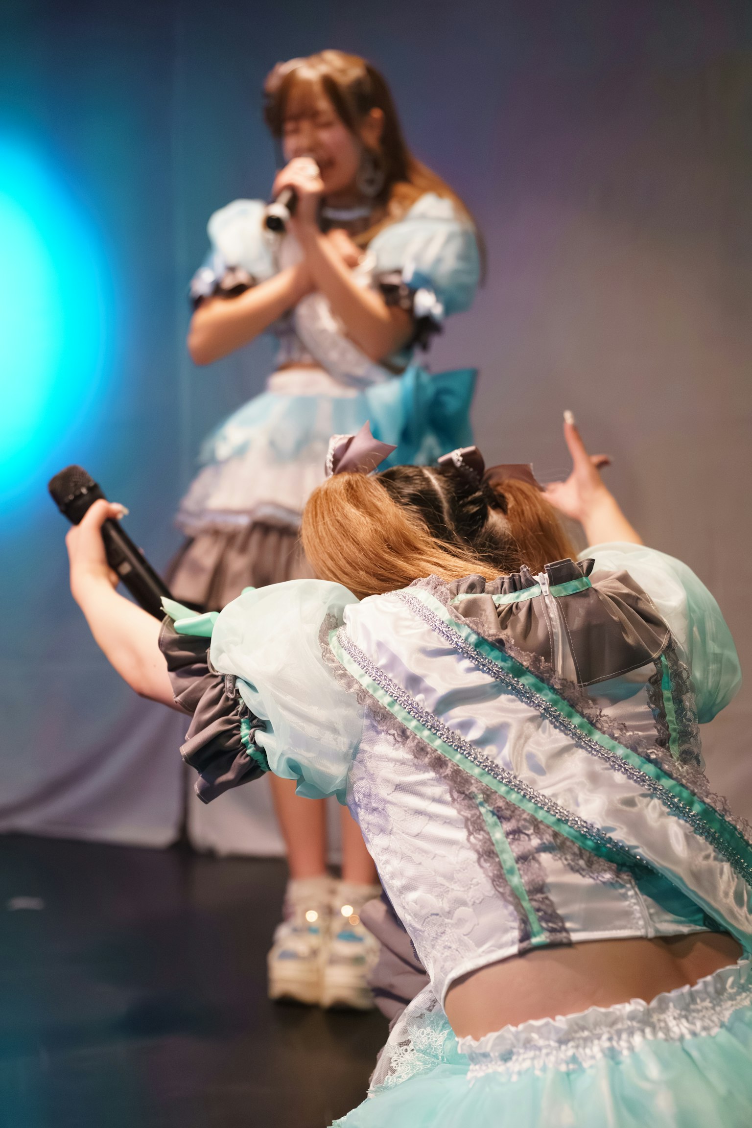 ステージでパフォーマンスするアイドルグループのメンバー二人が、青い衣装を着て歌っている