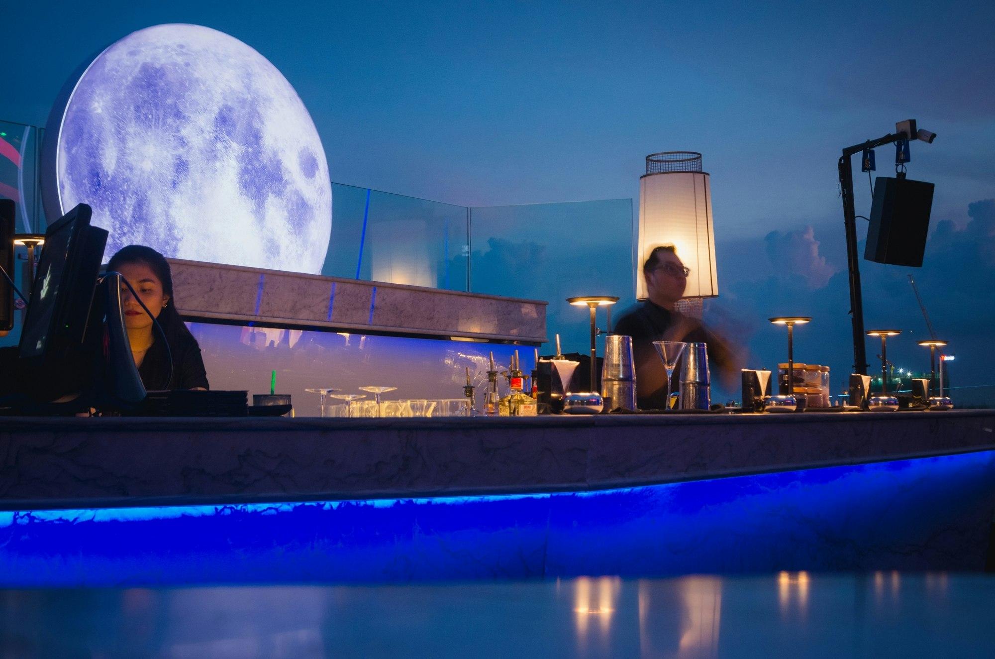 青いネオンのカウンター越しに月のような照明が浮かぶバーの夜景