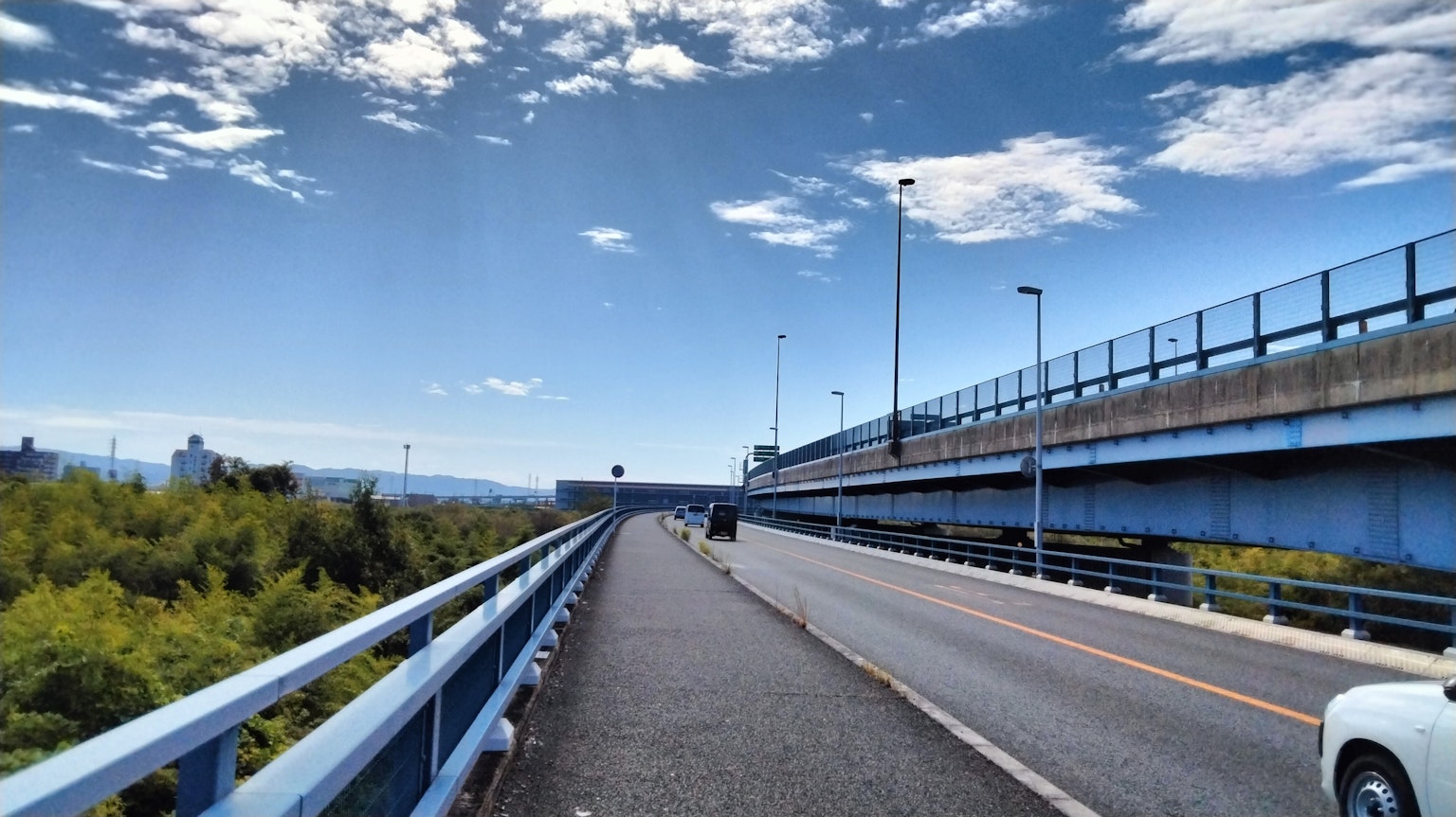 Scenic view of a blue bridge and road under a clear blue sky with clouds and green trees