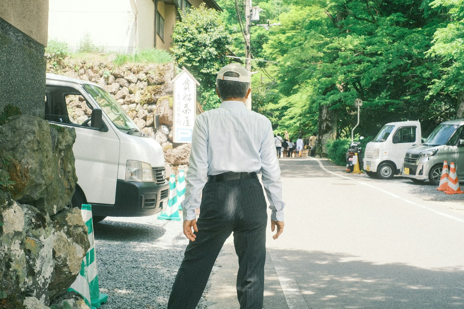ชายในเสื้อเชิ้ตสีขาวและกางเกงสีดำยืนอยู่บนถนนที่เงียบสงบรายล้อมไปด้วยต้นไม้สีเขียว