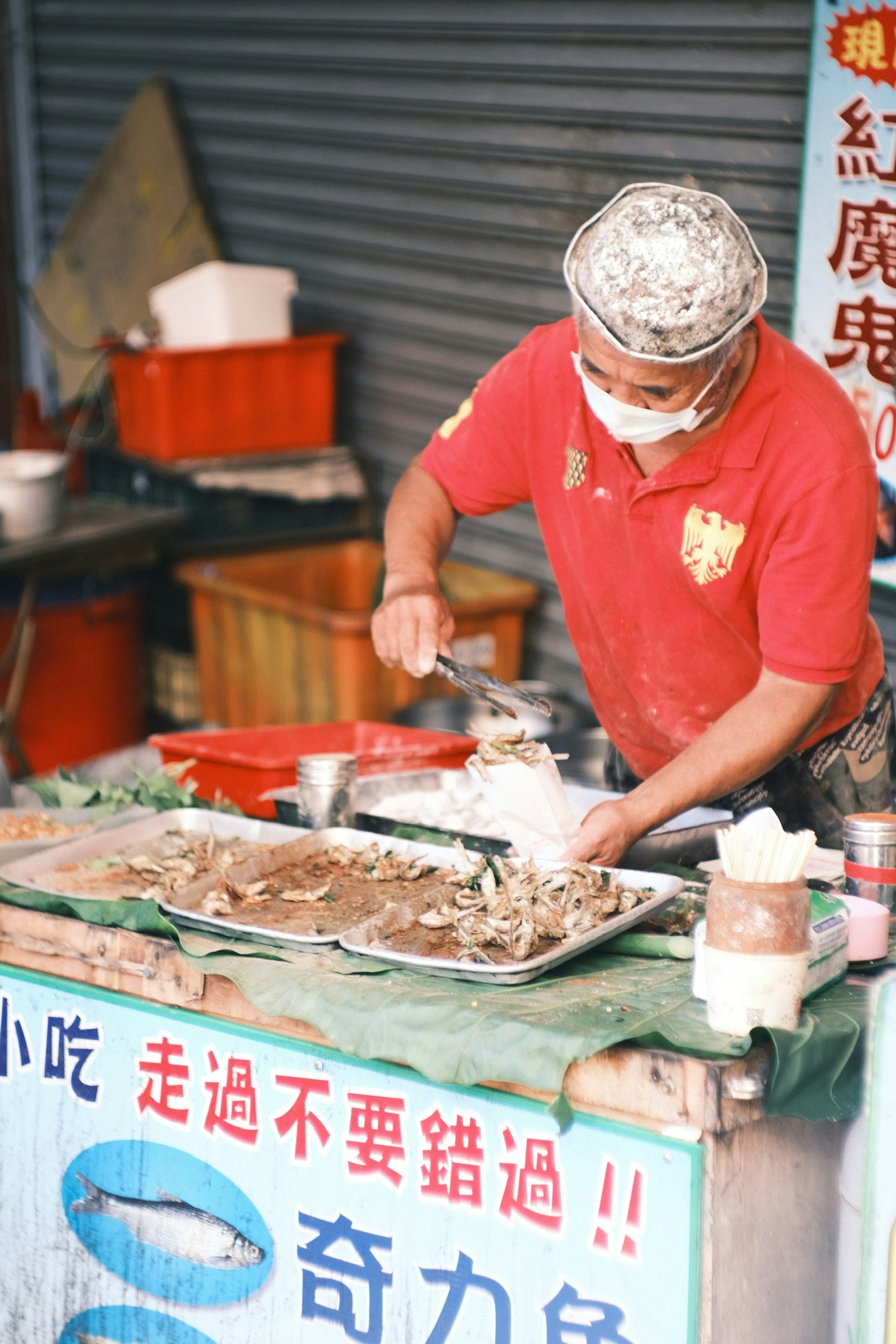 Hombre preparando comida en un puesto callejero con una camisa roja y una máscara