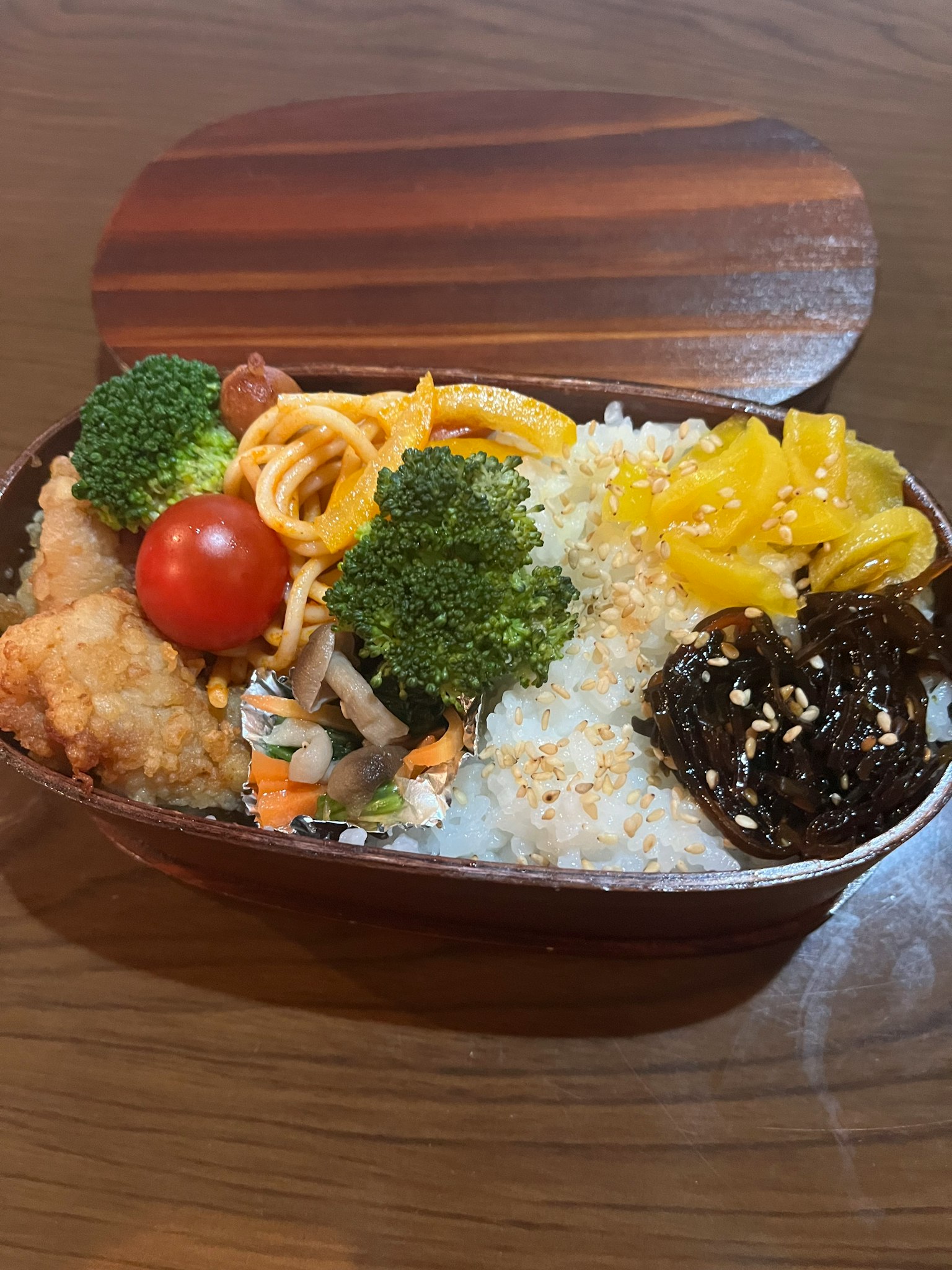Una caja bento llena de arroz, brócoli, tomates cherry, espagueti, pollo frito, alga, mango