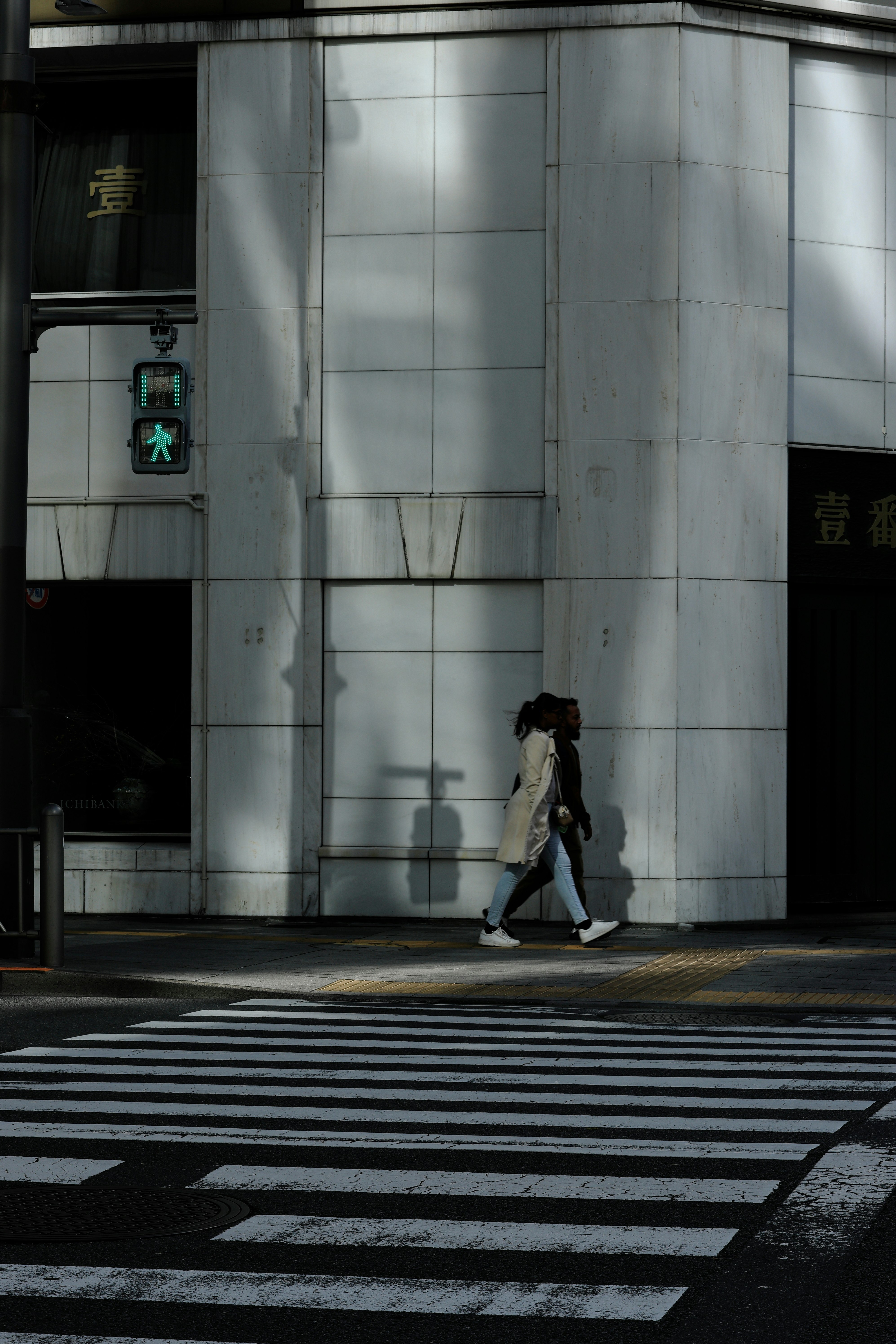 白い壁の前を歩く二人の人物と影のある交差点