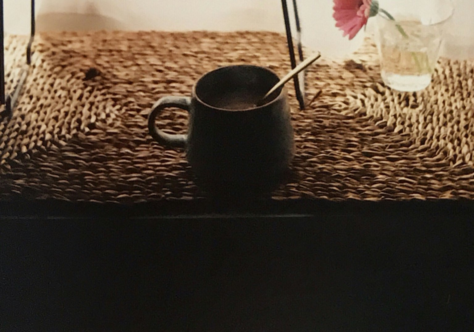 Una tazza nera con un cucchiaio su una superficie intrecciata accanto a un fiore in un bicchiere