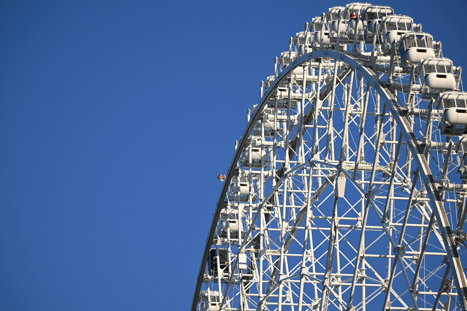 Part of a large ferris wheel against a blue sky featuring white structure and capsules