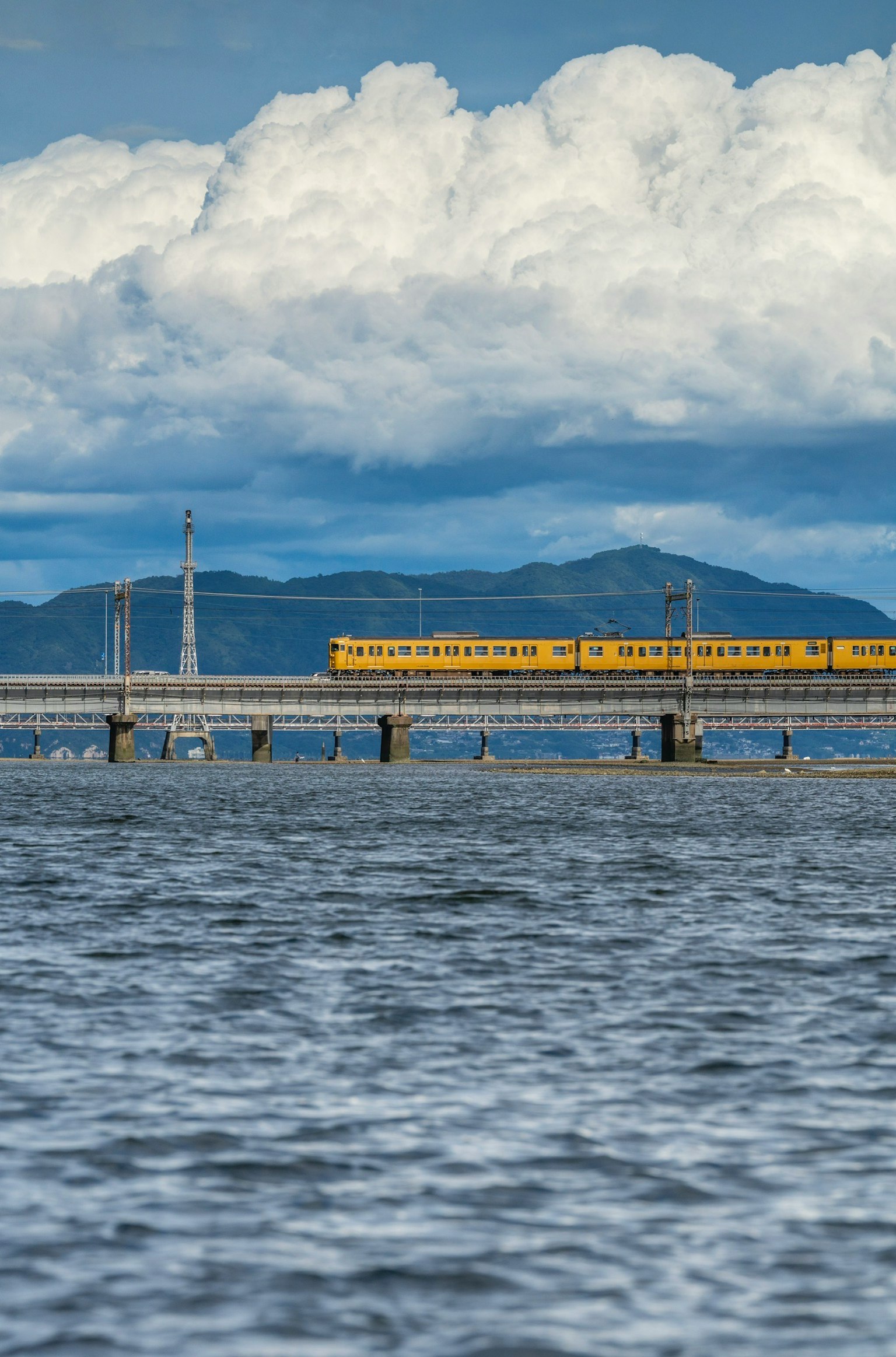 A yellow train crossing a bridge over blue water with clouds in the sky