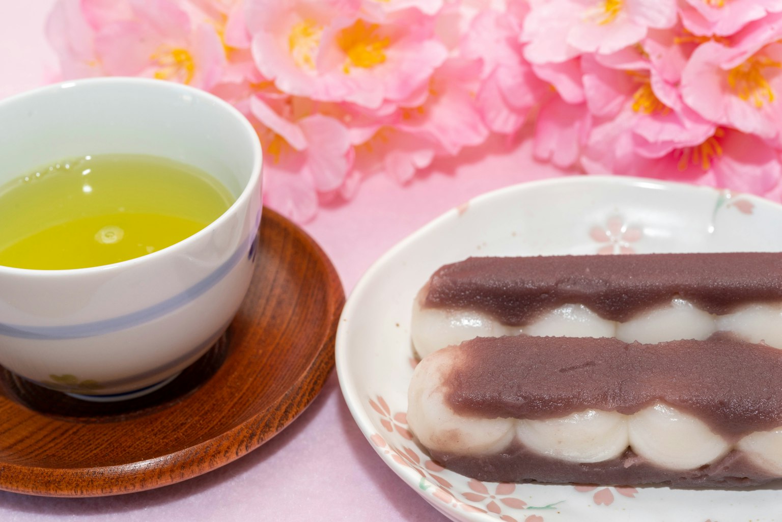 Secangkir teh hijau di samping makanan manis tradisional Jepang di atas piring dengan bunga merah muda di latar belakang