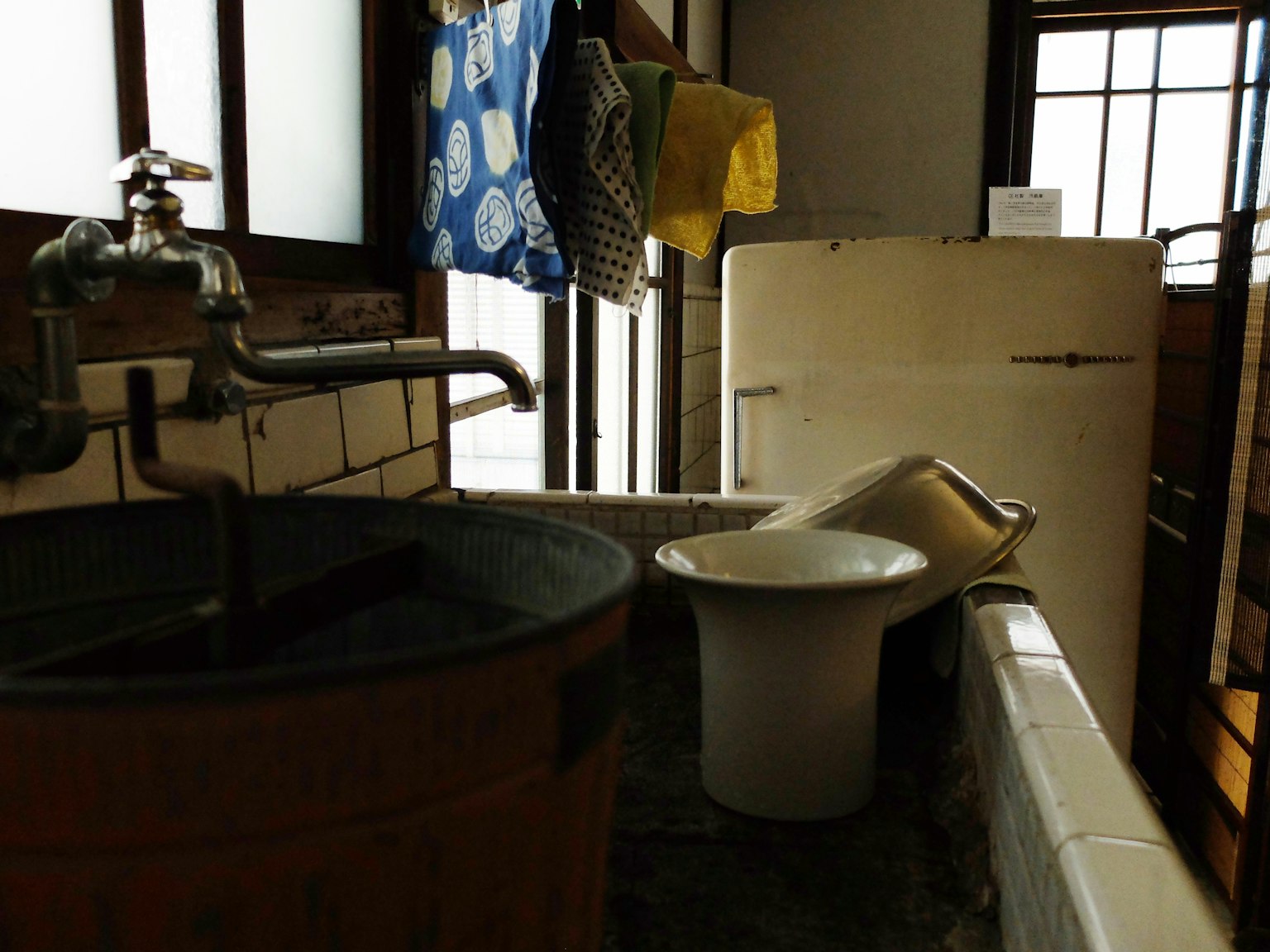 Interno di un vecchio bagno giapponese con una vasca di legno e un WC in ceramica, luce naturale che entra dalle finestre e biancheria stesa