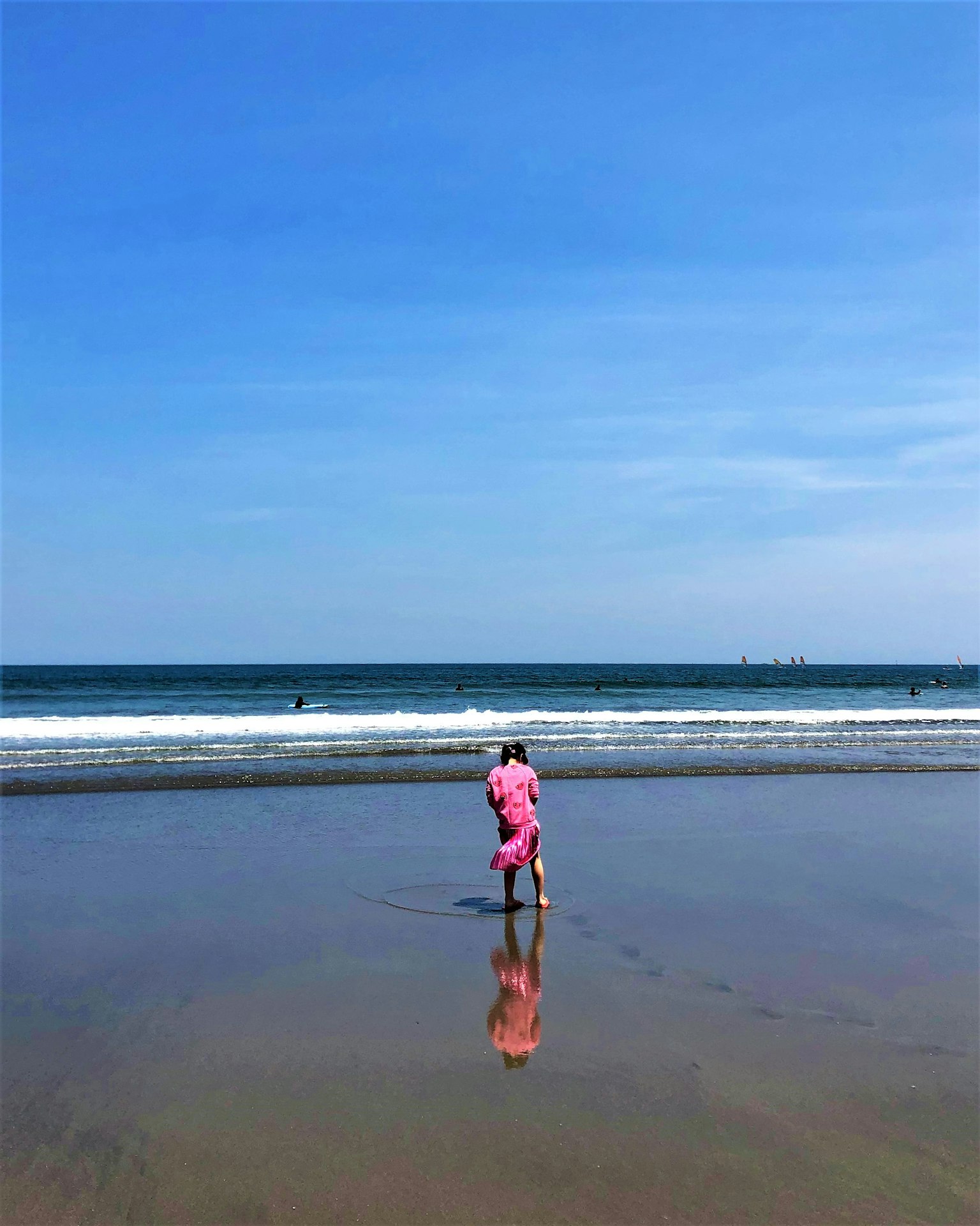 A child in a pink outfit walking along the shoreline with waves and a blue sky