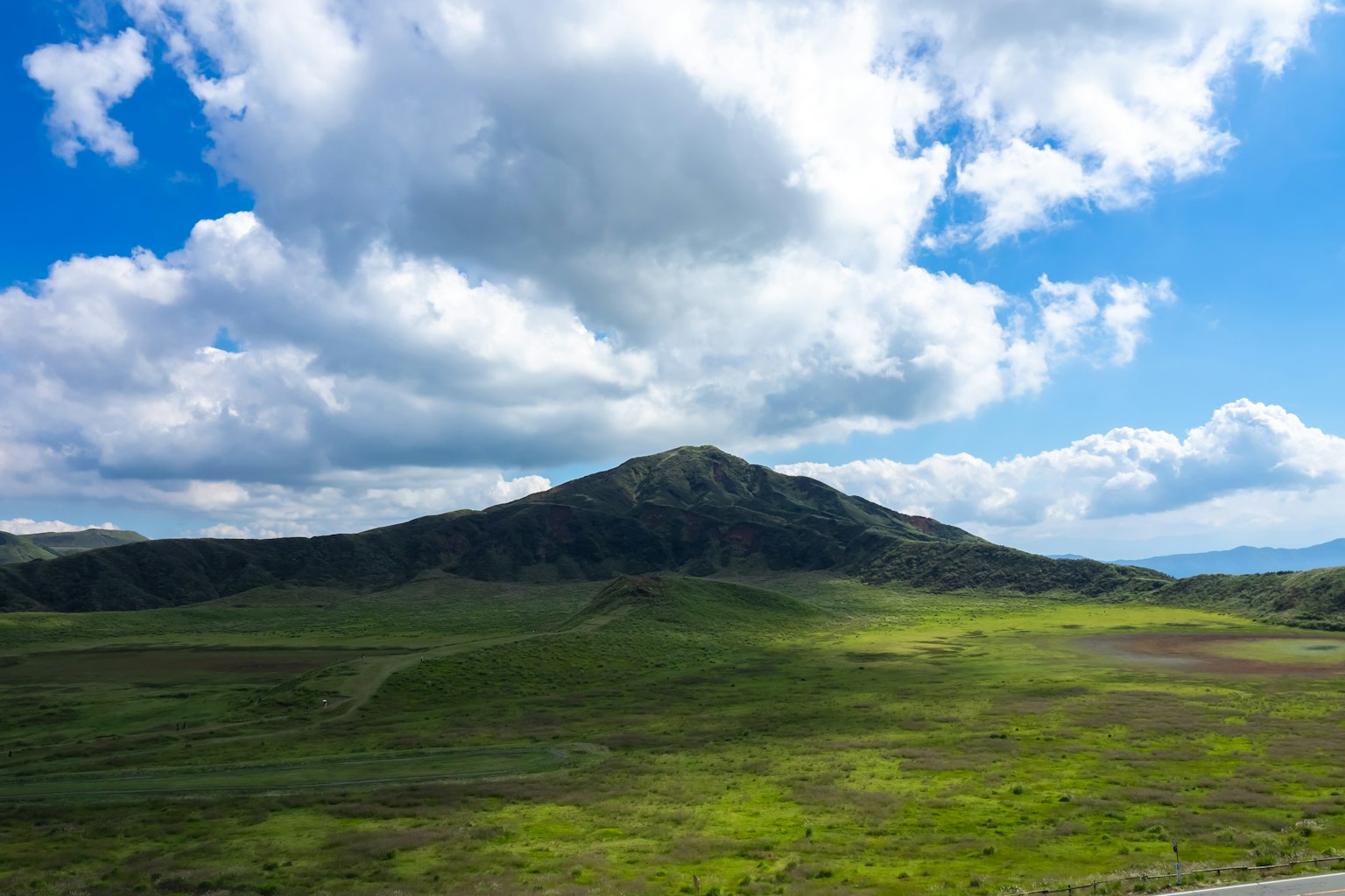 Una montaña que se eleva sobre un prado verde rodeado de cielo azul y nubes blancas