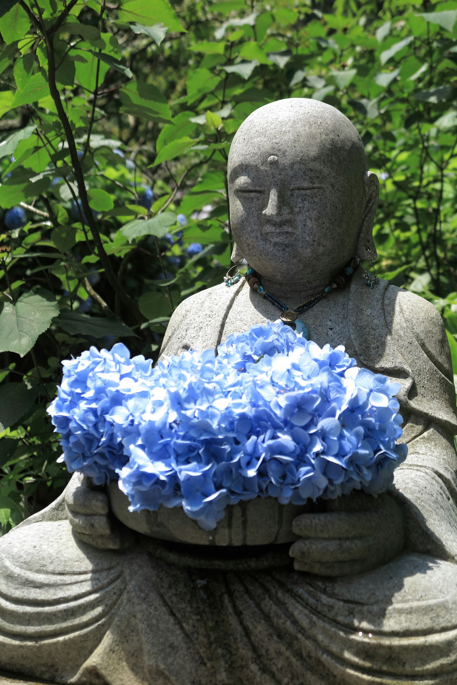 Statue de Bouddha en pierre tenant un bouquet de fleurs bleues entourée de verdure