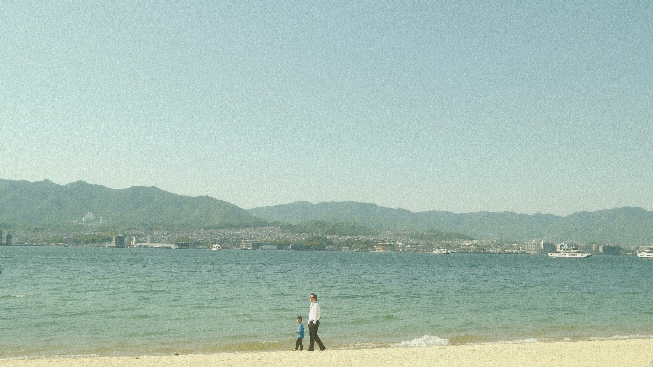 穏やかな海辺と遠くの山並みを眺める人物のいる砂浜
