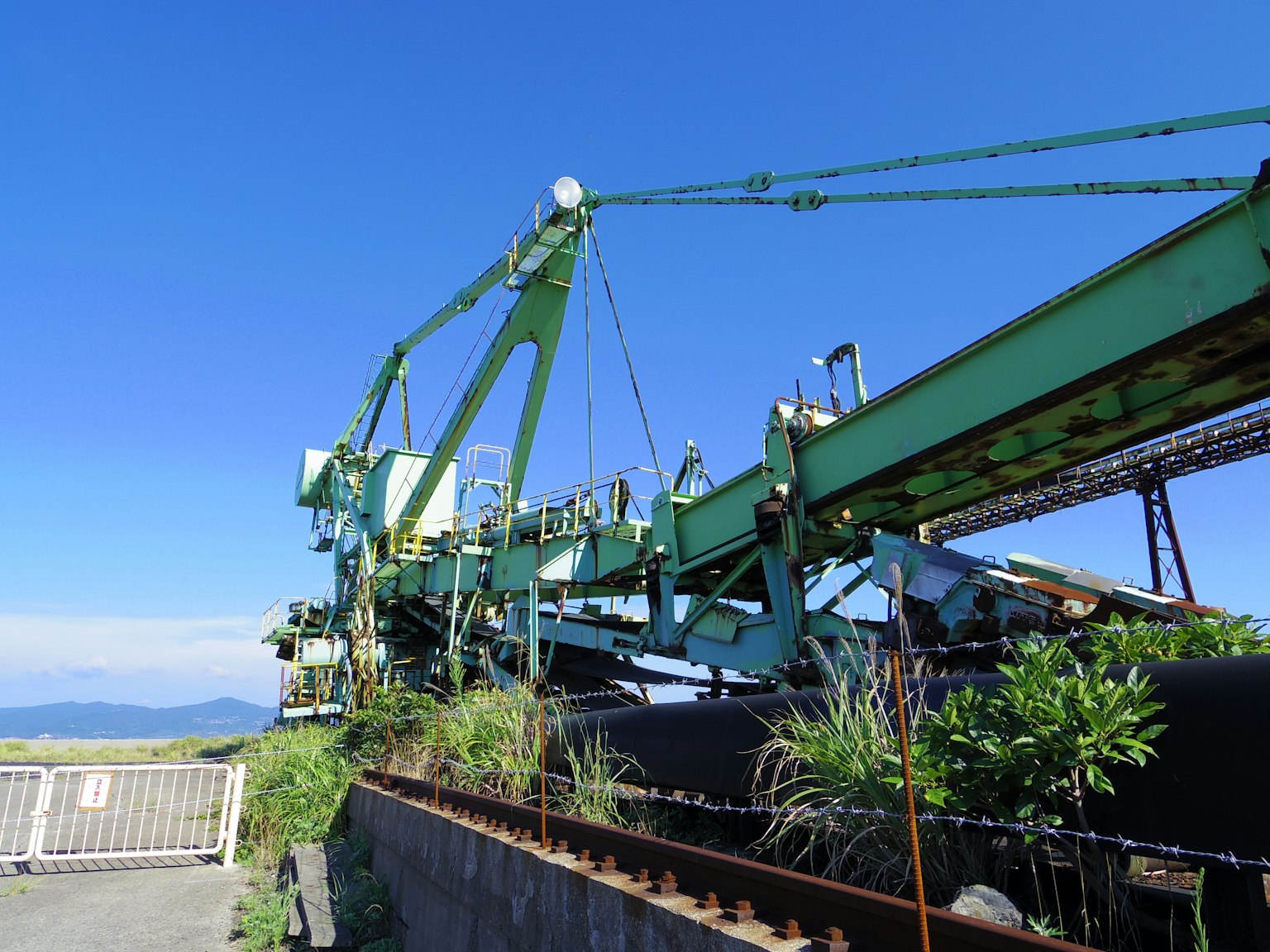 โครงสร้างเครนสีเขียวและสายพานลำเลียงใต้ท้องฟ้าสีฟ้าพร้อมพืชที่เติบโตใกล้เคียง