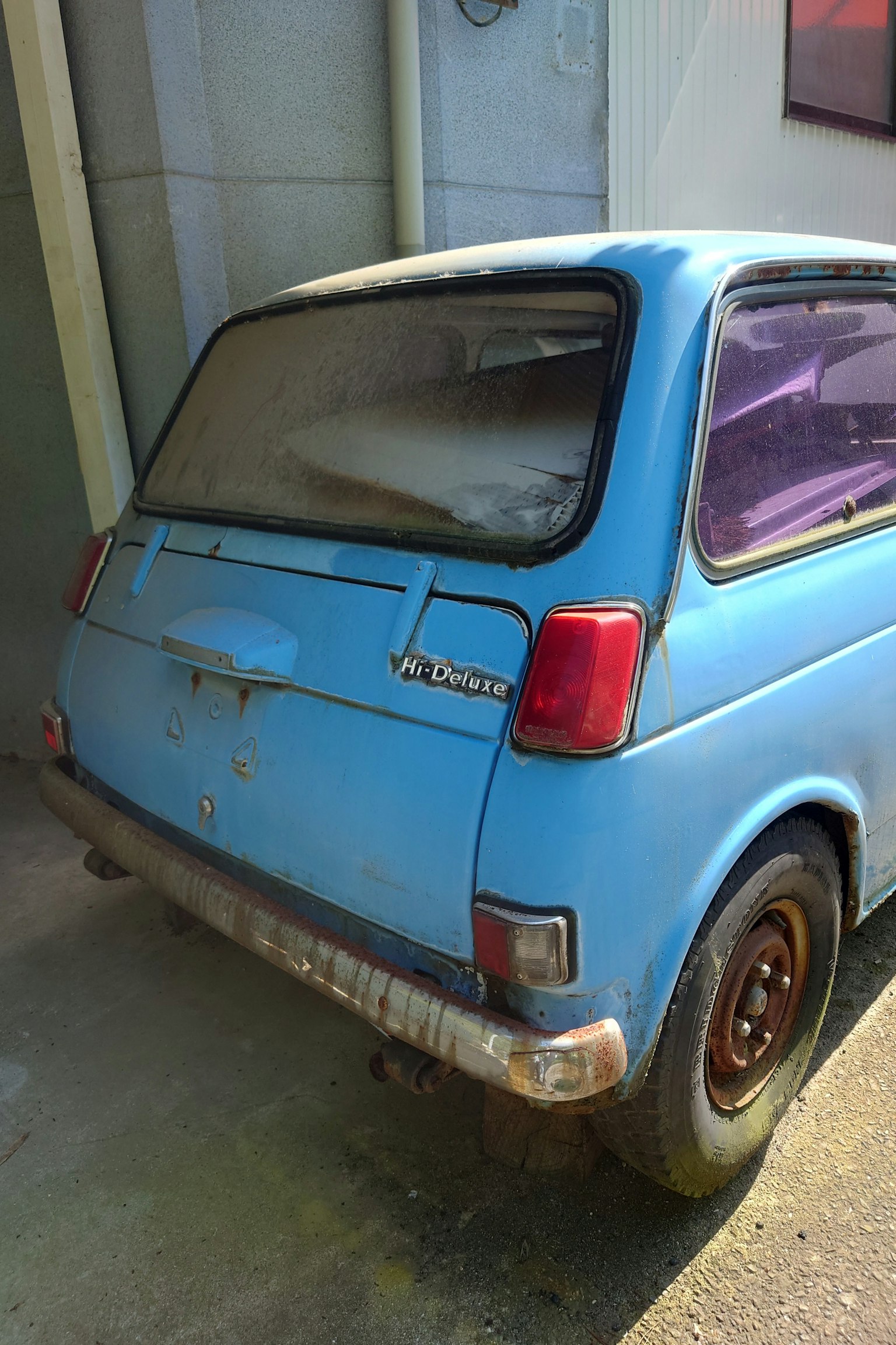 Vista trasera de un viejo coche compacto azul con una ventana polvorienta y carrocería oxidada