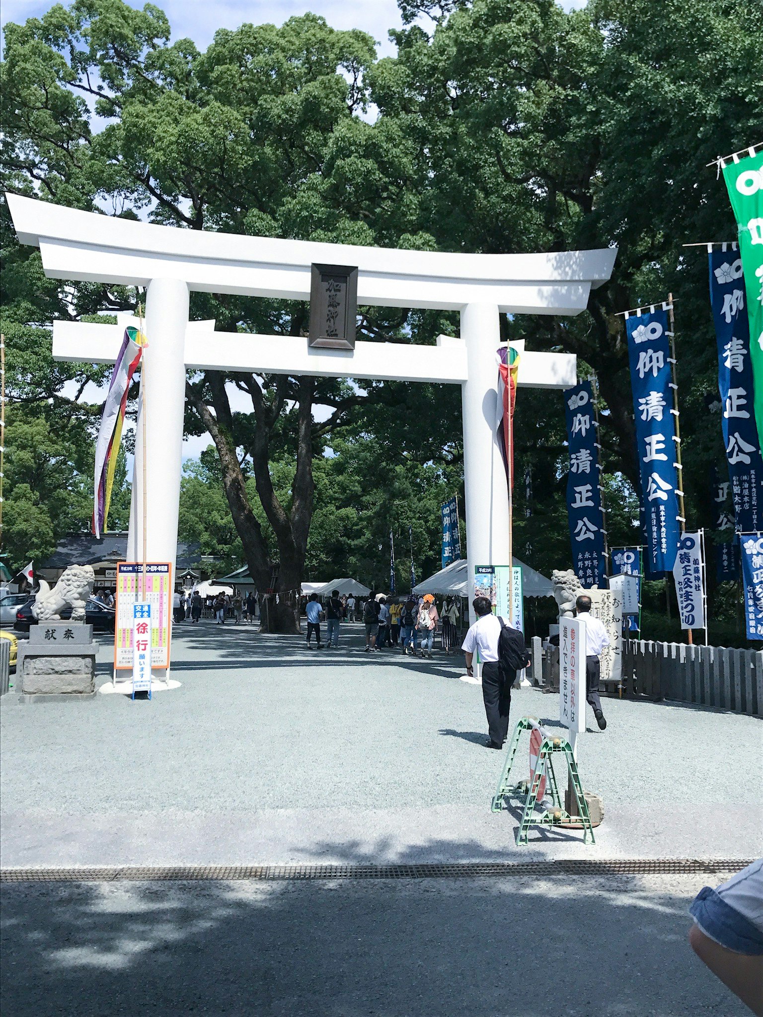 Entrance to a shrine featuring a white torii gate with people walking by surrounded by lush greenery