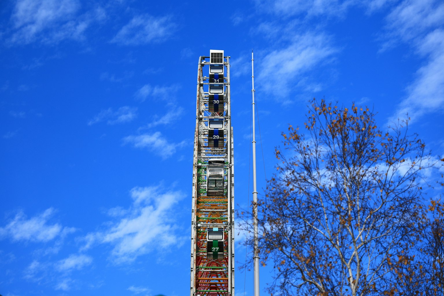 Bunte Riesenradstruktur unter einem blauen Himmel mit Wolken und einem Teil eines Baumes