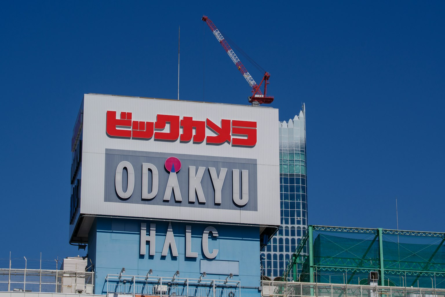 Gran letrero con 'ODAKYU' y 'Bic Camera' en un edificio bajo un cielo azul claro con una grúa visible