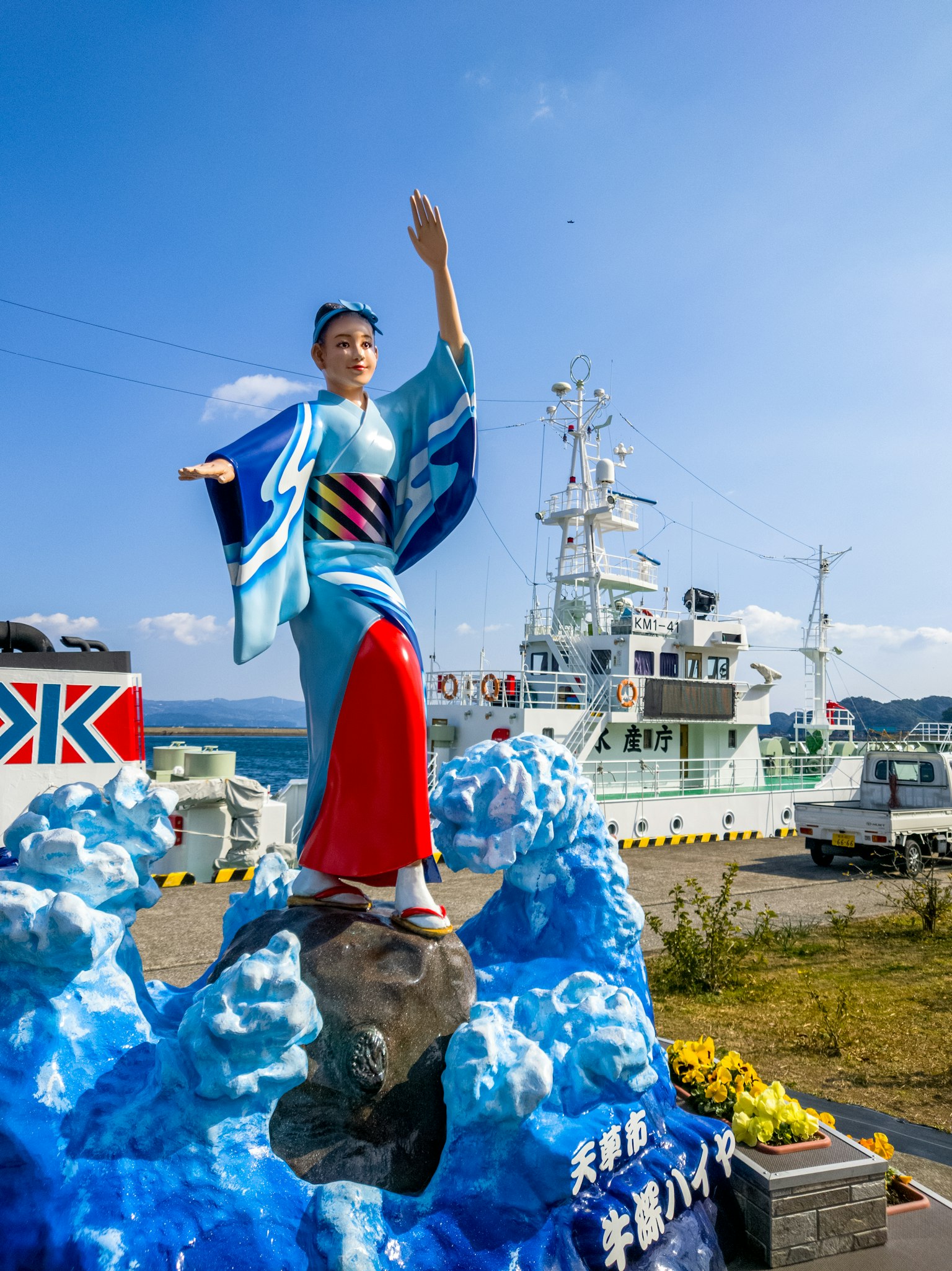 穿著和服的女性雕像站在藍色波浪上，背景中有一艘船
