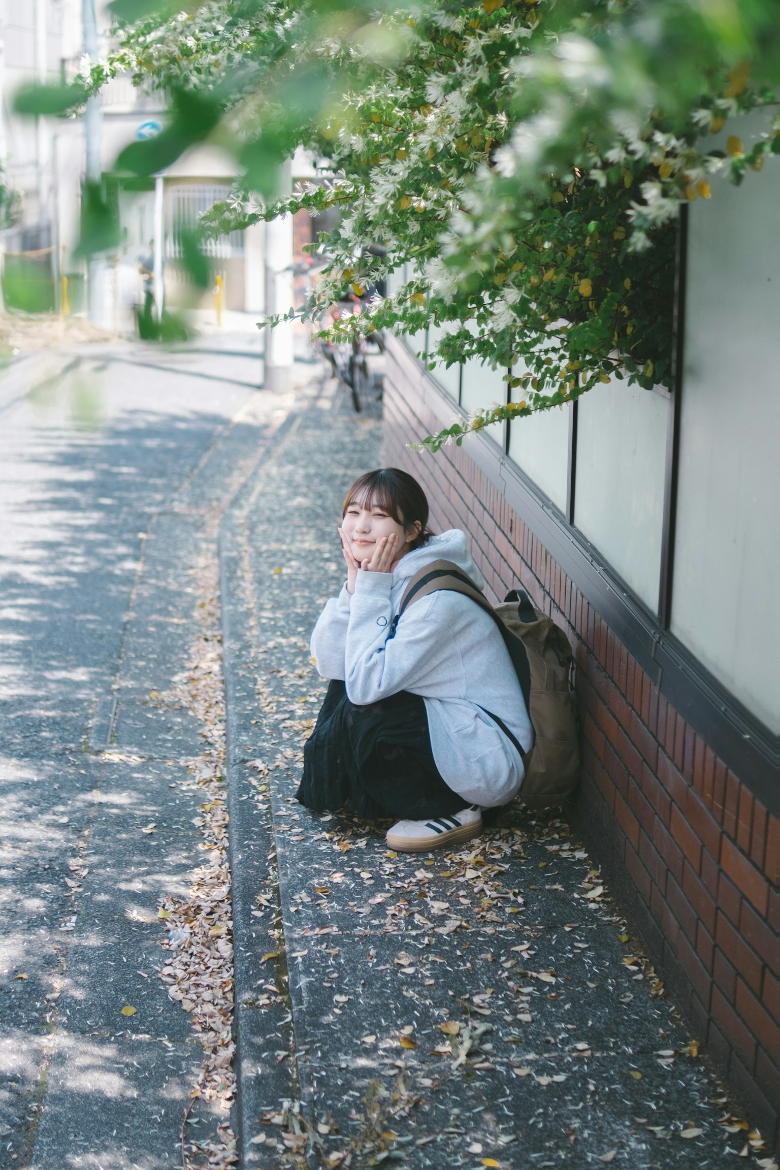 A woman sitting by the roadside wearing a gray hoodie surrounded by fallen leaves and greenery