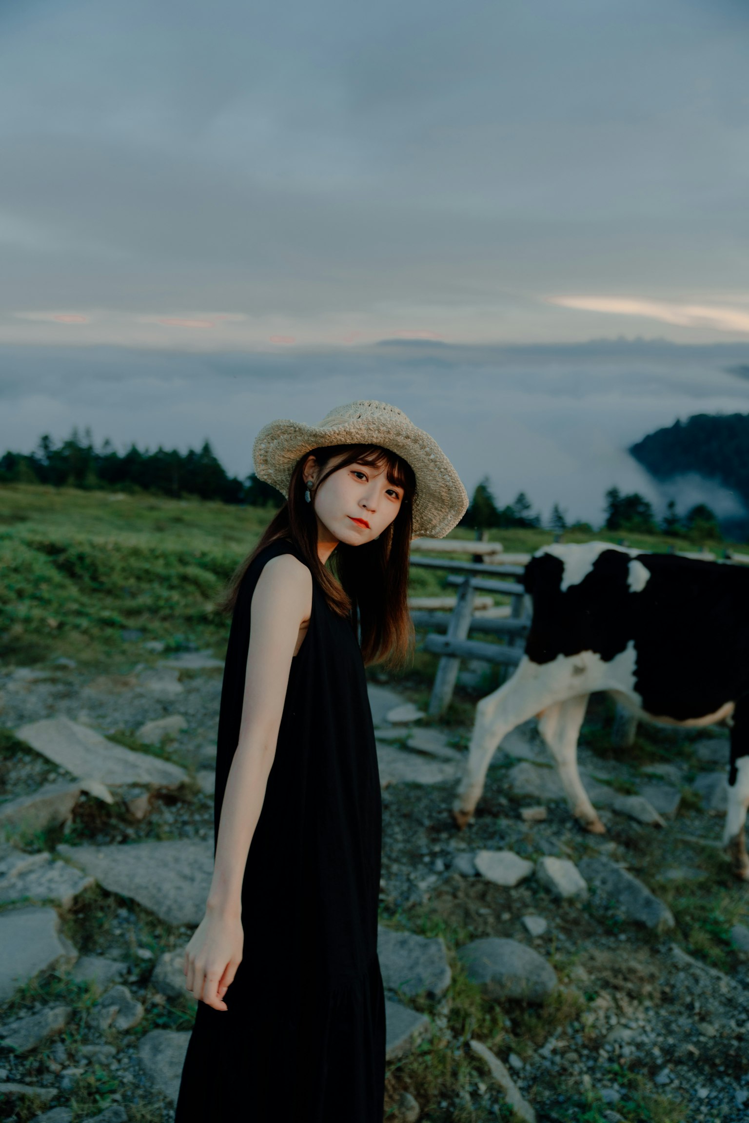 Una donna in abito nero con un cappello di paglia in piedi vicino a una mucca in un paesaggio pittoresco