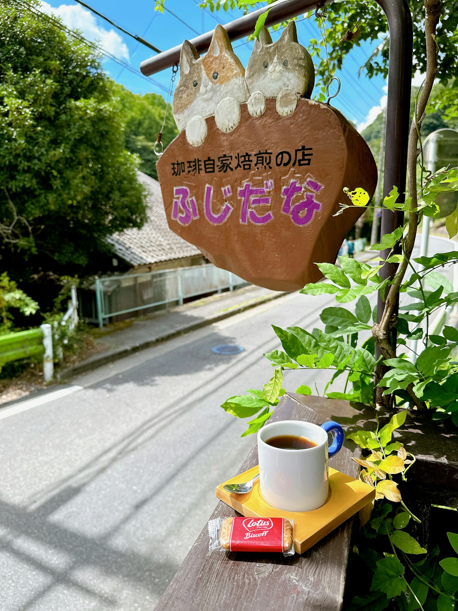 小咖啡館桌上的咖啡和小吃 木製招牌上寫著藤田名字 綠色植物和藍天背景
