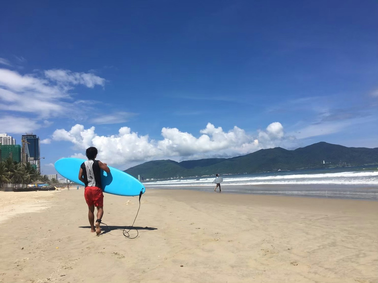Man walking on the beach holding a blue surfboard with a clear sky and ocean in the background