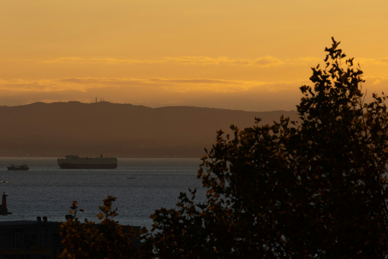 Vista del atardecer sobre el mar con montañas al fondo barcos navegando en el agua y árboles en silueta