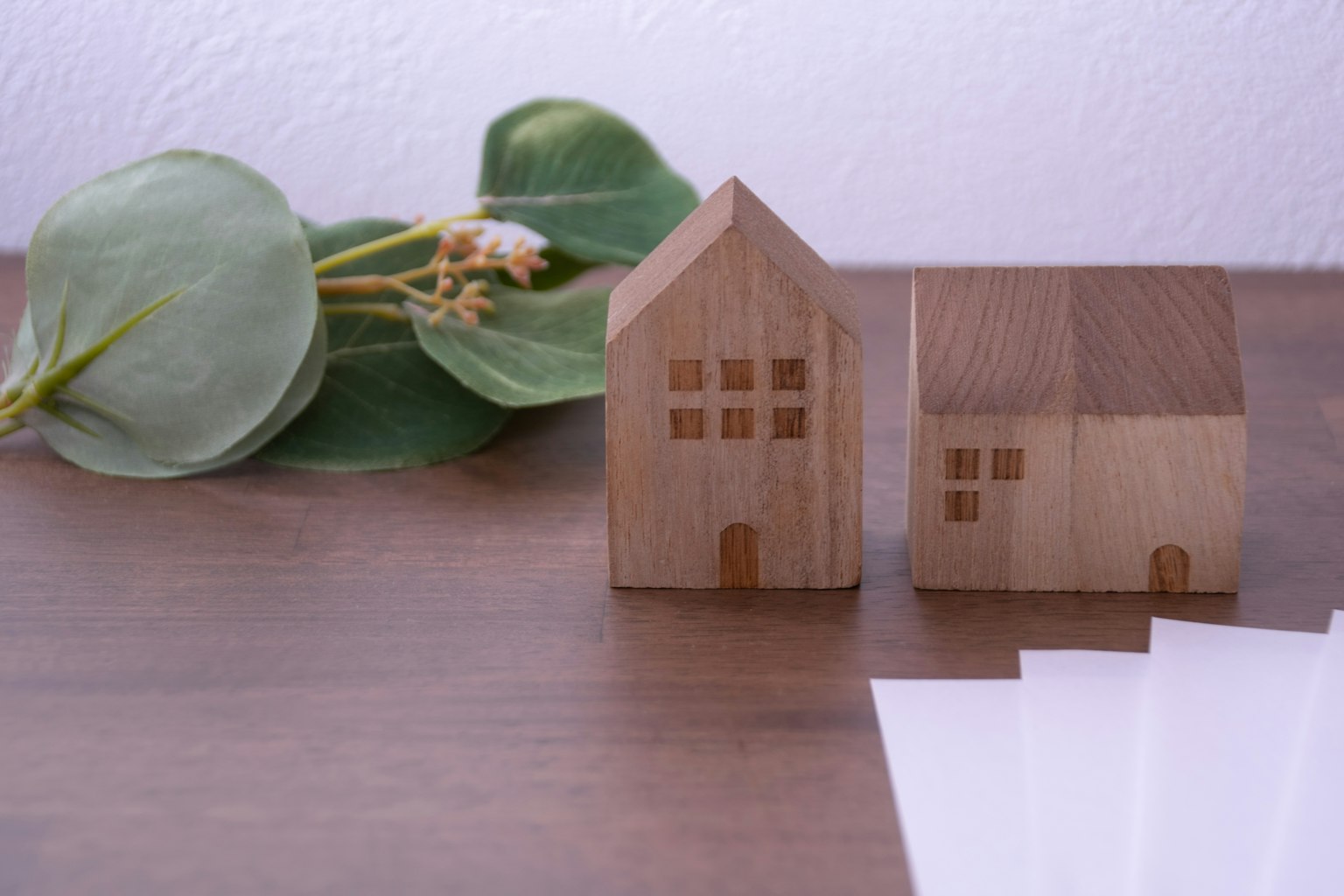 Two wooden house models placed on a wooden surface with green leaves and white paper nearby