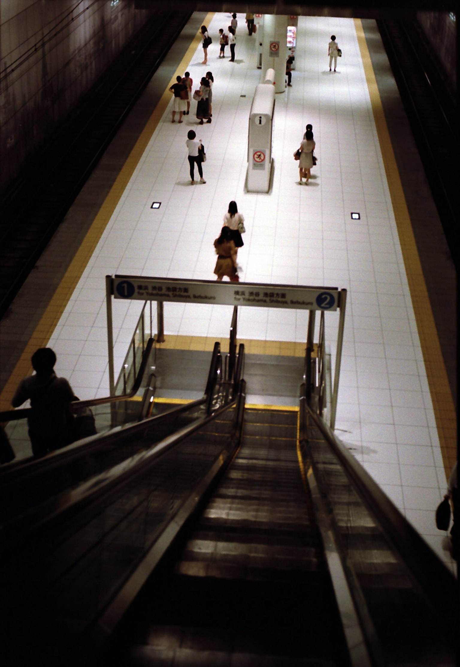 Vue depuis un escalier roulant dans une station de métro des personnes marchant sur un chemin carrelé lumineux