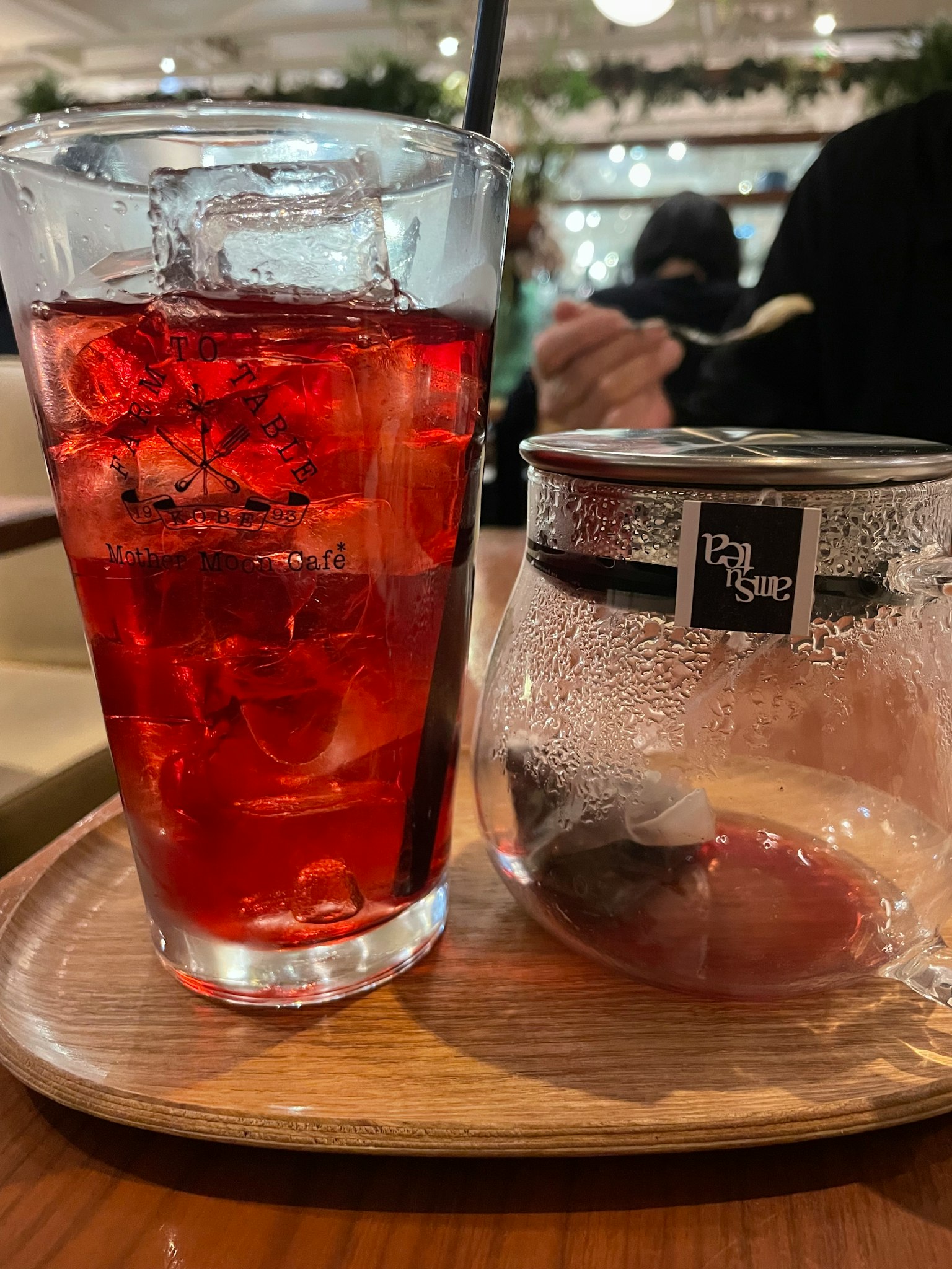 A glass of red drink with ice on a wooden tray next to a small jar