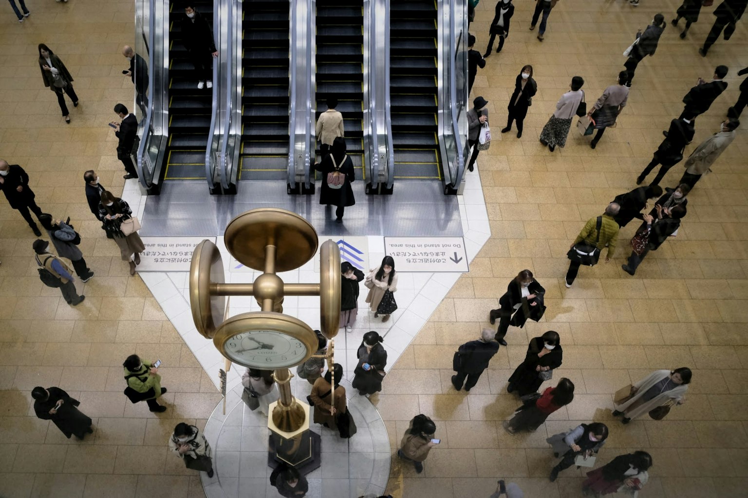Một quảng trường đông đúc với thang cuốn và nhiều người di chuyển quanh một chiếc đồng hồ trung tâm