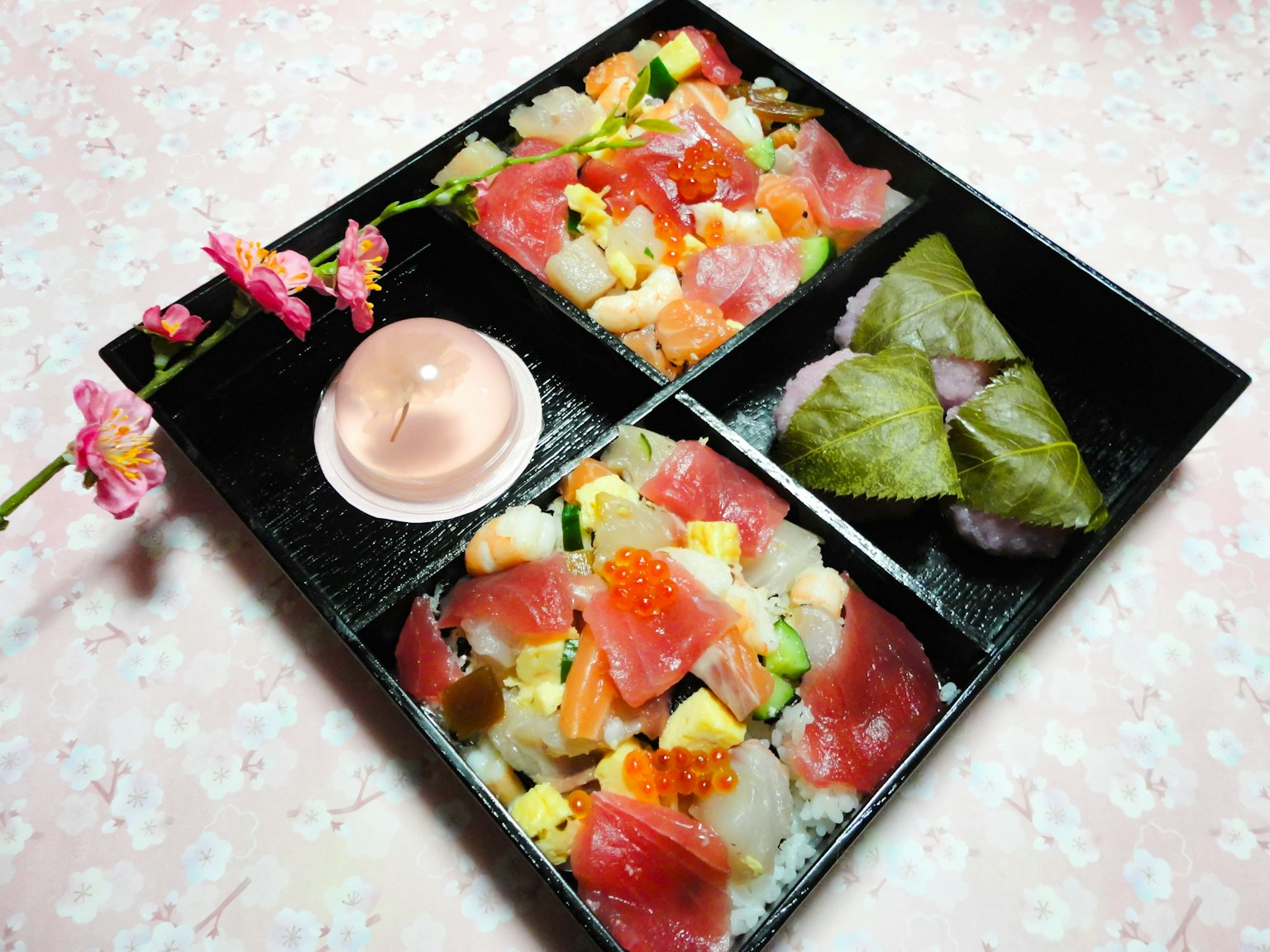 Bento de sushi bellamente presentado con pescado colorido y verduras en cuatro secciones acompañado de una flor de cerezo y una salsa