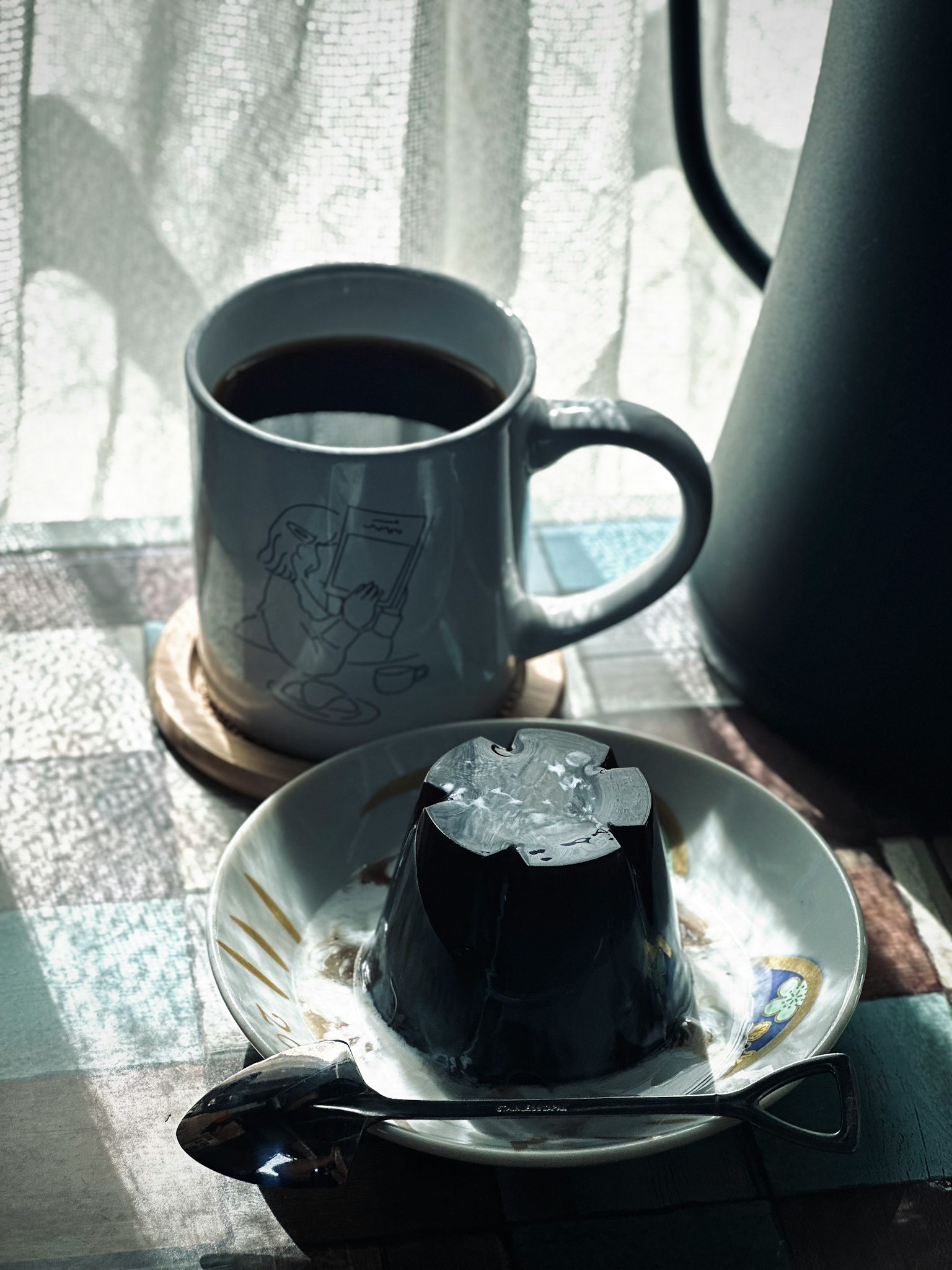 Una escena serena con café y postre una taza blanca llena de café una gelatina negra con un diseño floral cerca de una ventana con luz solar