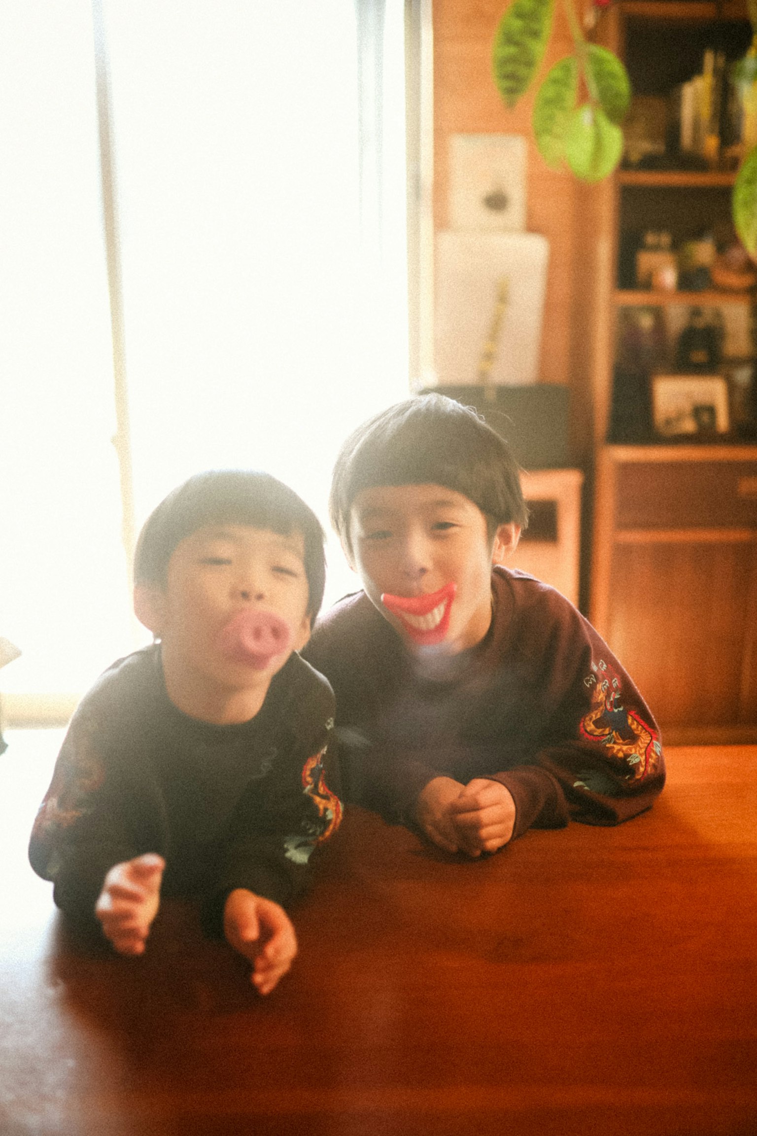二人の子供がテーブルの前で遊んでいる 笑顔で面白い表情をしている 明るい室内の光が差し込んでいる